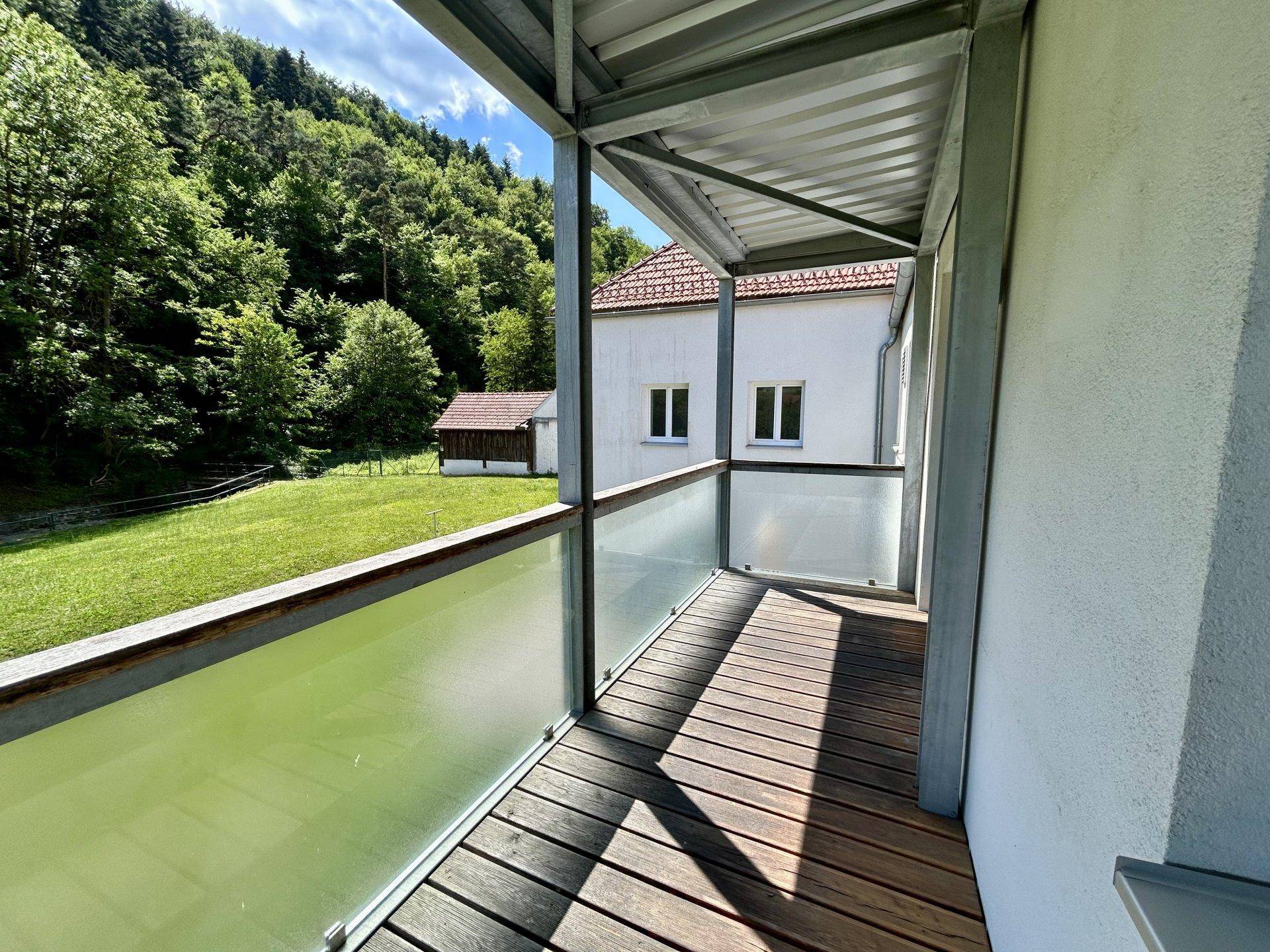 Balkon mit Holzboden und Glasgeländer, umgeben von grüner Natur und Gebäuden.