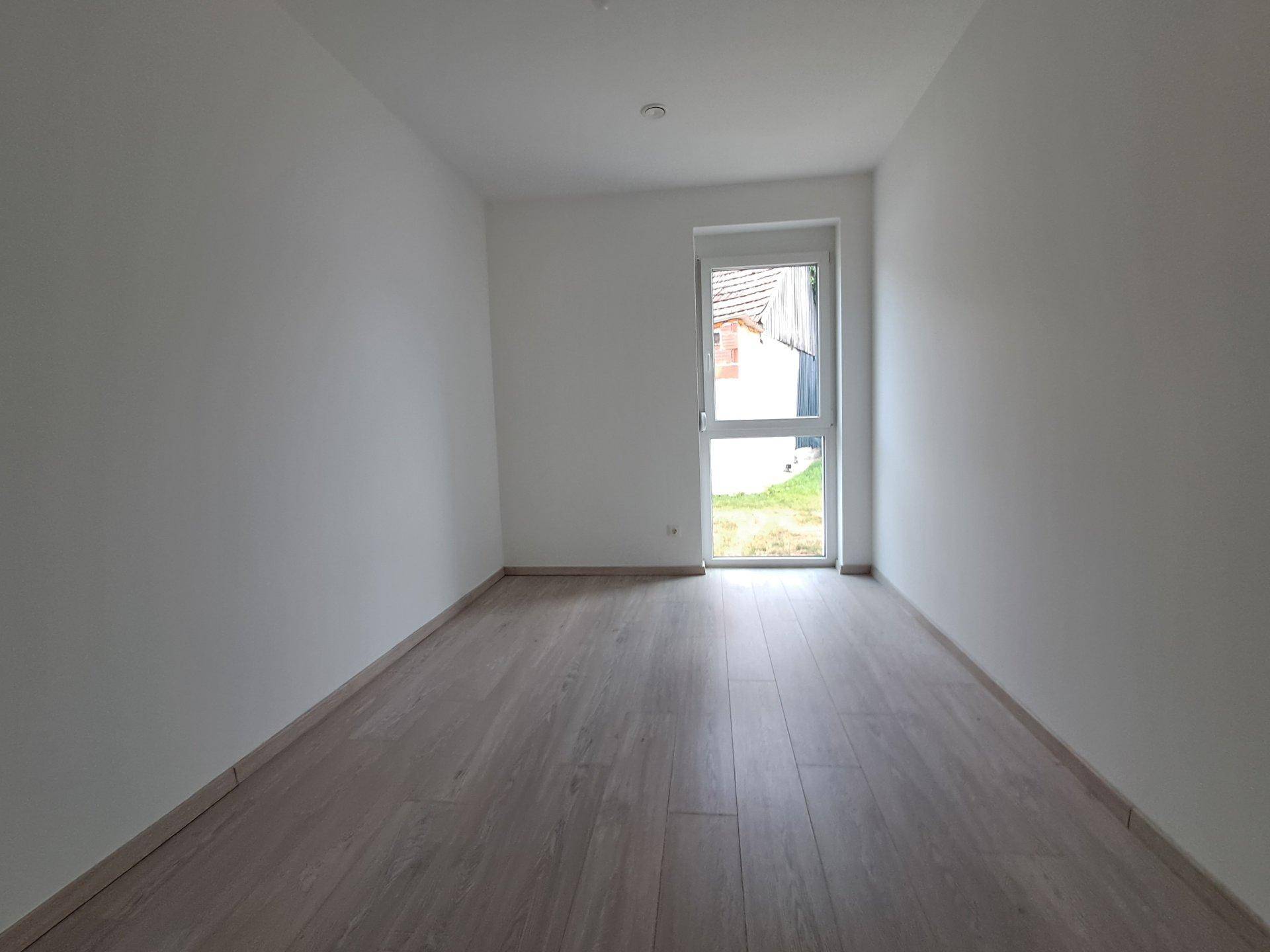 Helles, leeres Zimmer mit Laminatboden und einem Fenster, das den Blick ins Grüne freigibt.