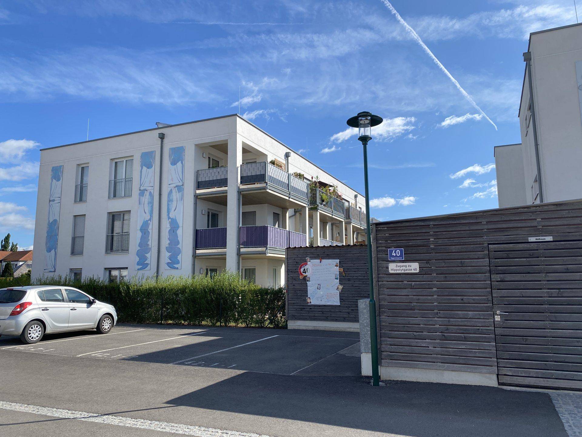 Modernes Apartmentgebäude mit Balkonen und weißer Fassade, umgeben von Grünflächen und Parkplätzen.