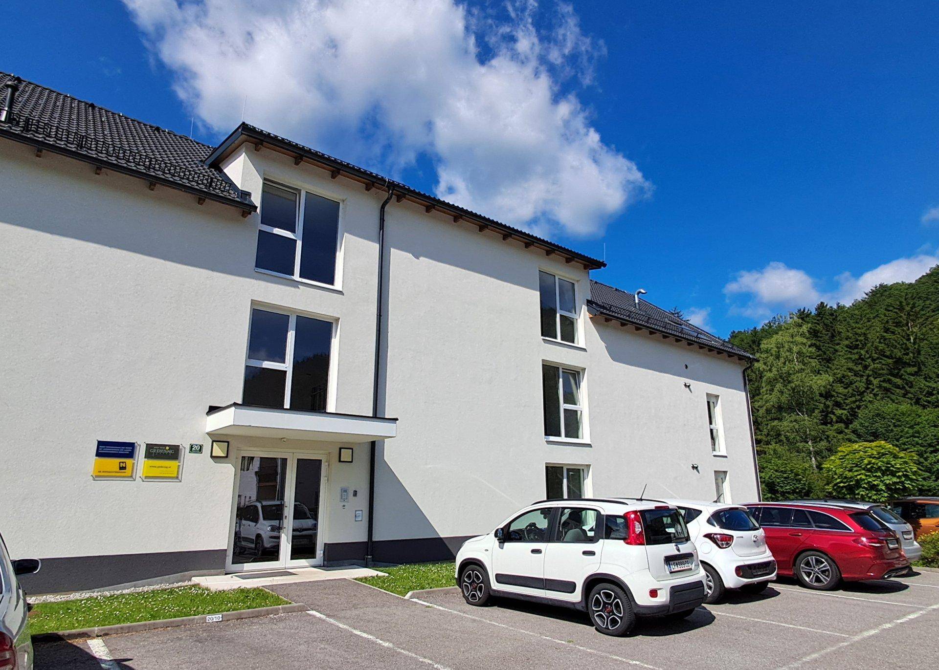 Haupteingang des Mehrfamilienhauses mit davorliegenden Parkplätzen und grüner Umgebung.