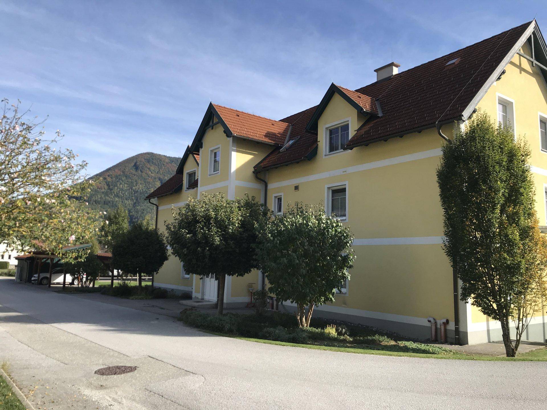 Gelbes Wohngebäude mit rotem Ziegeldach und grünen Bäumen in ländlicher Umgebung.