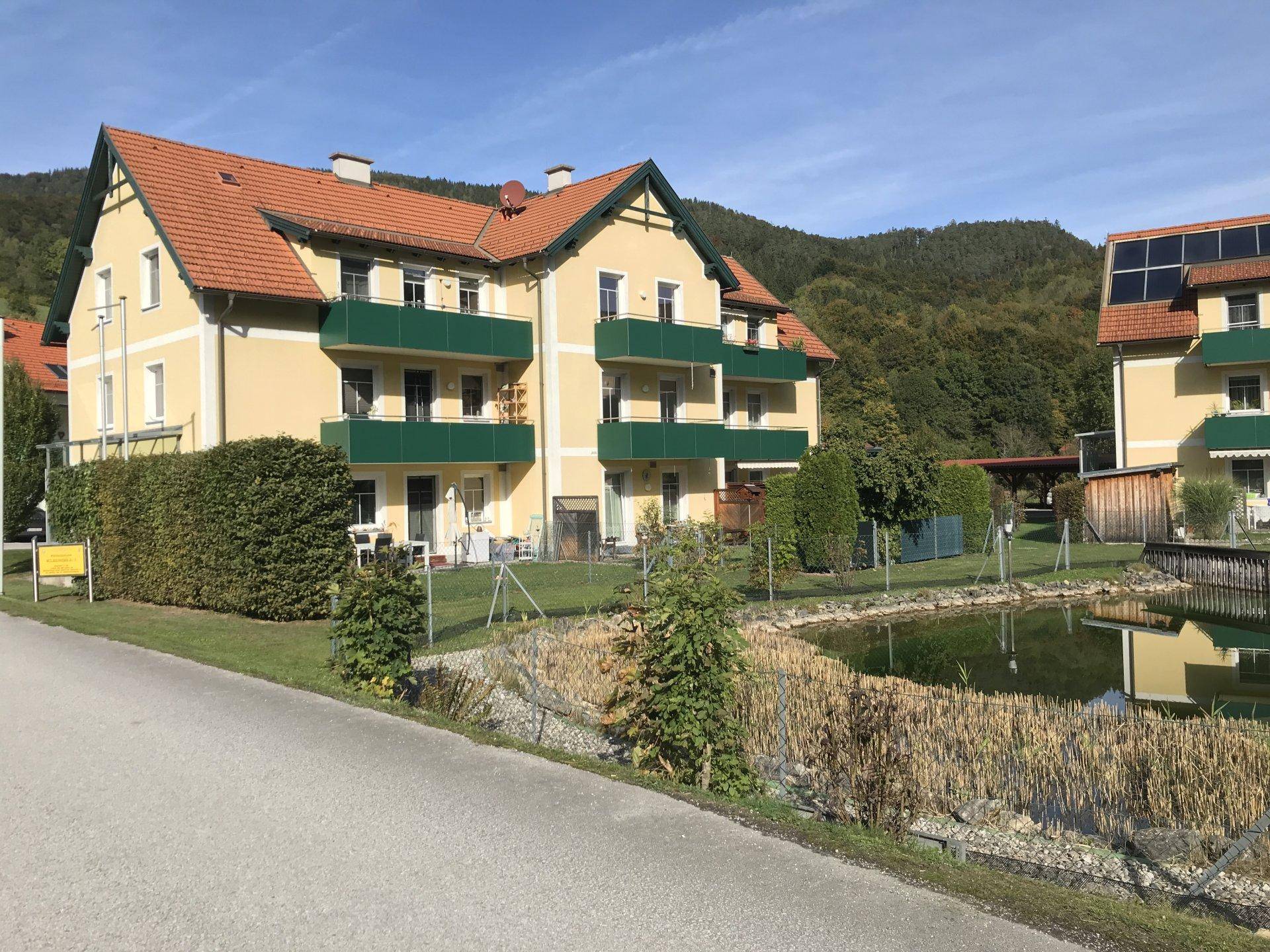Wohngebäude mit Balkonen und einem Naturschwimmteich, umgeben von grüner Natur und Bergen.