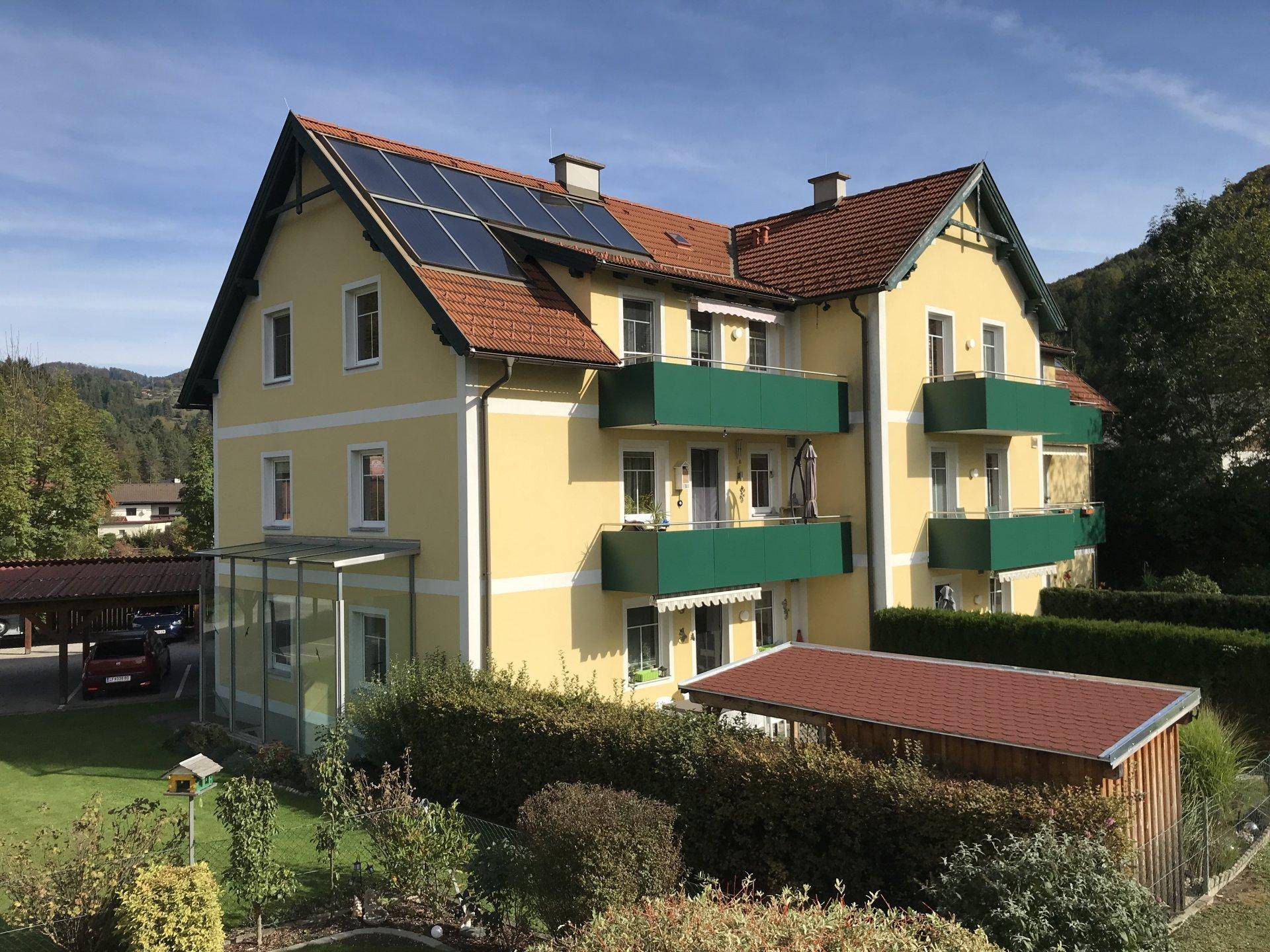 Gelbes Wohnhaus mit grünen Balkonen, Solaranlagen auf dem Dach und einem Gartenhaus.