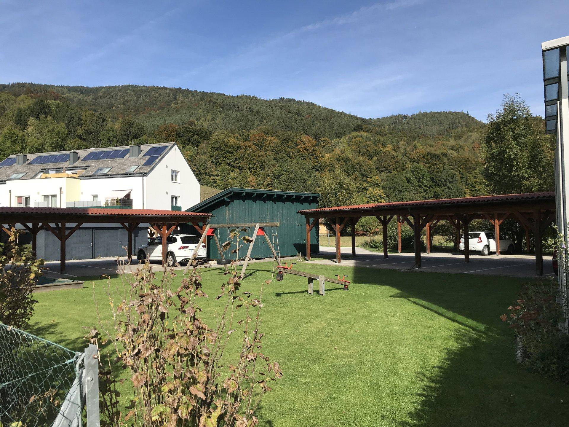 Grüner Spielplatz mit Schaukeln und Rutsche, umgeben von Carports und bewaldeten Bergen.