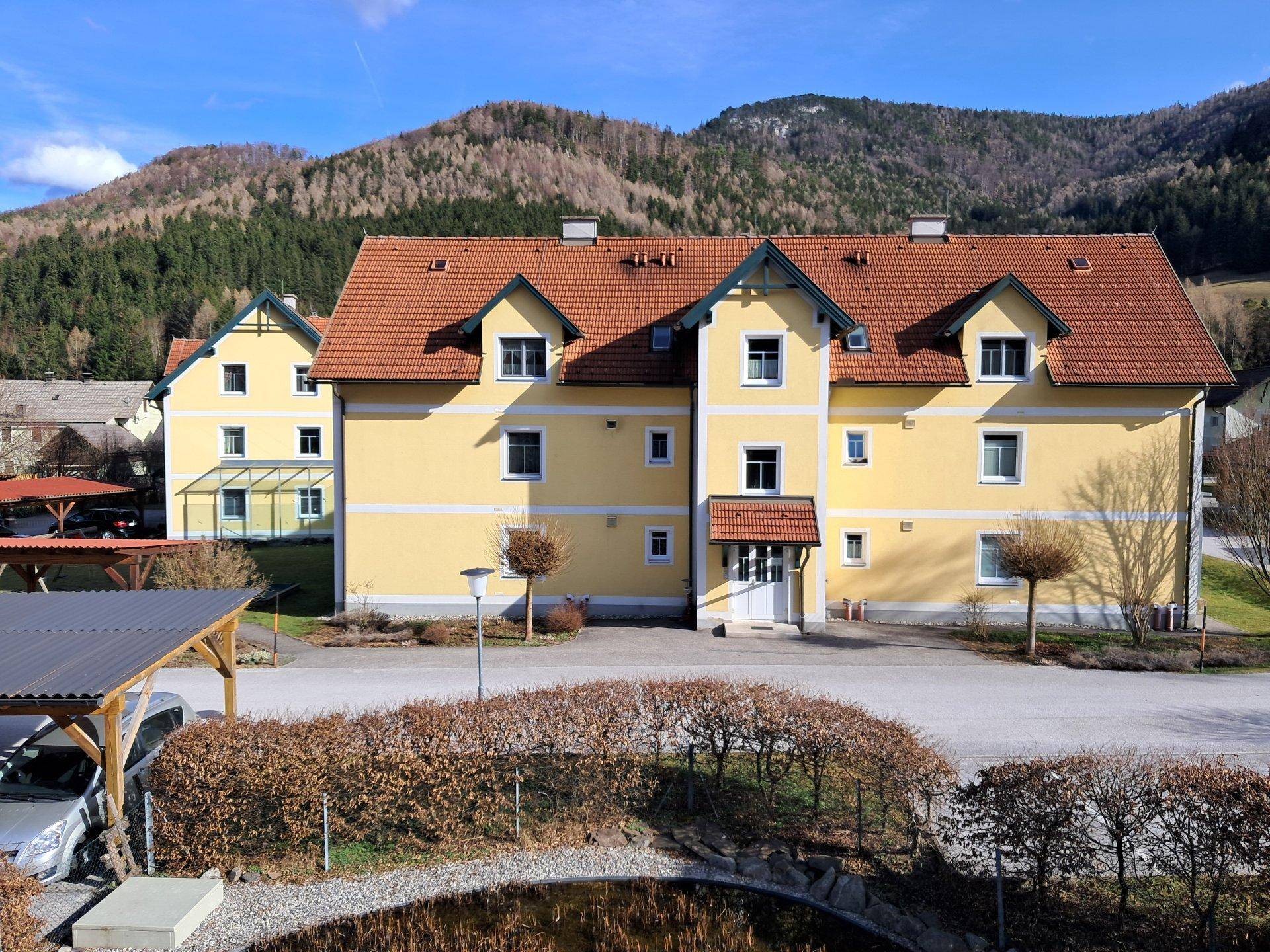 Gelbes Wohngebäude mit rotem Ziegeldach und mehreren Fenstern, umgeben von Bergen und Natur.