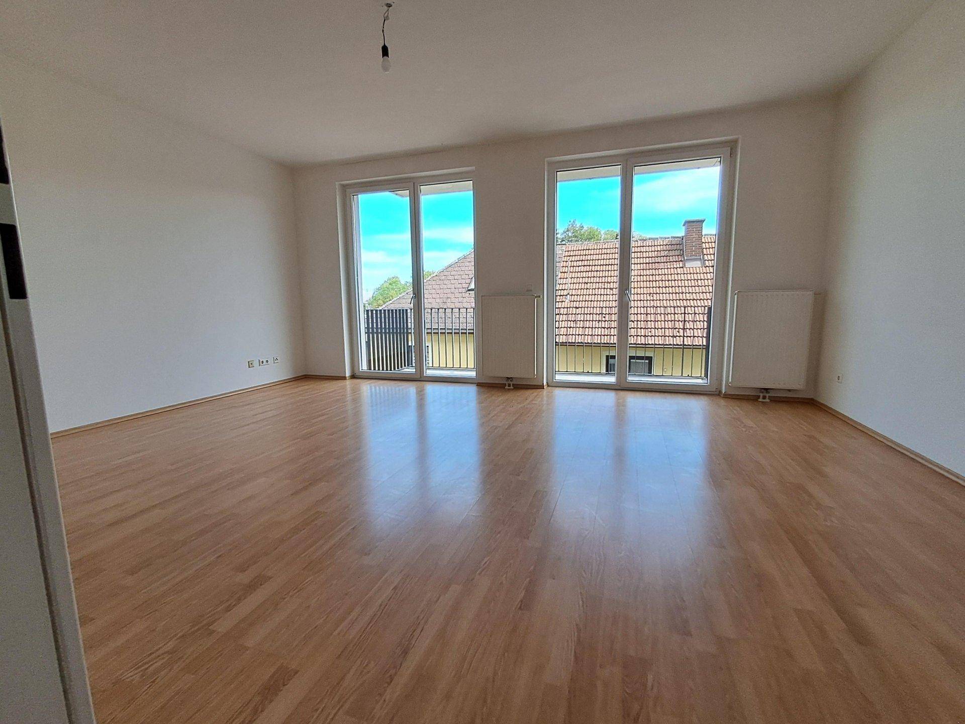Heller Wohnraum mit Laminatboden und großen Fenstern, die viel Tageslicht hereinlassen.