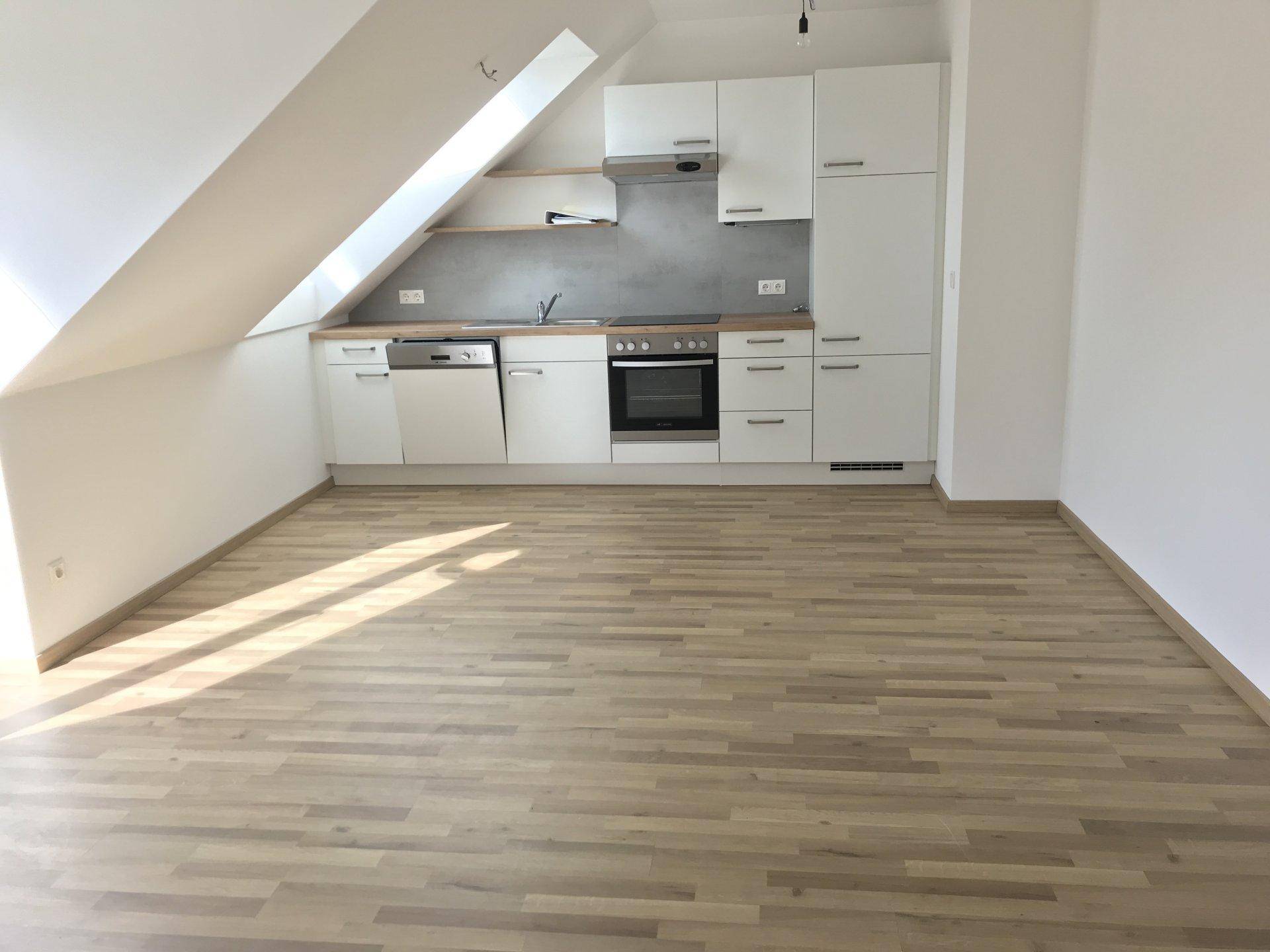 Offener Wohnbereich mit integrierter weißer Küche, Laminatboden und schrägen Wänden im Dachgeschoss.