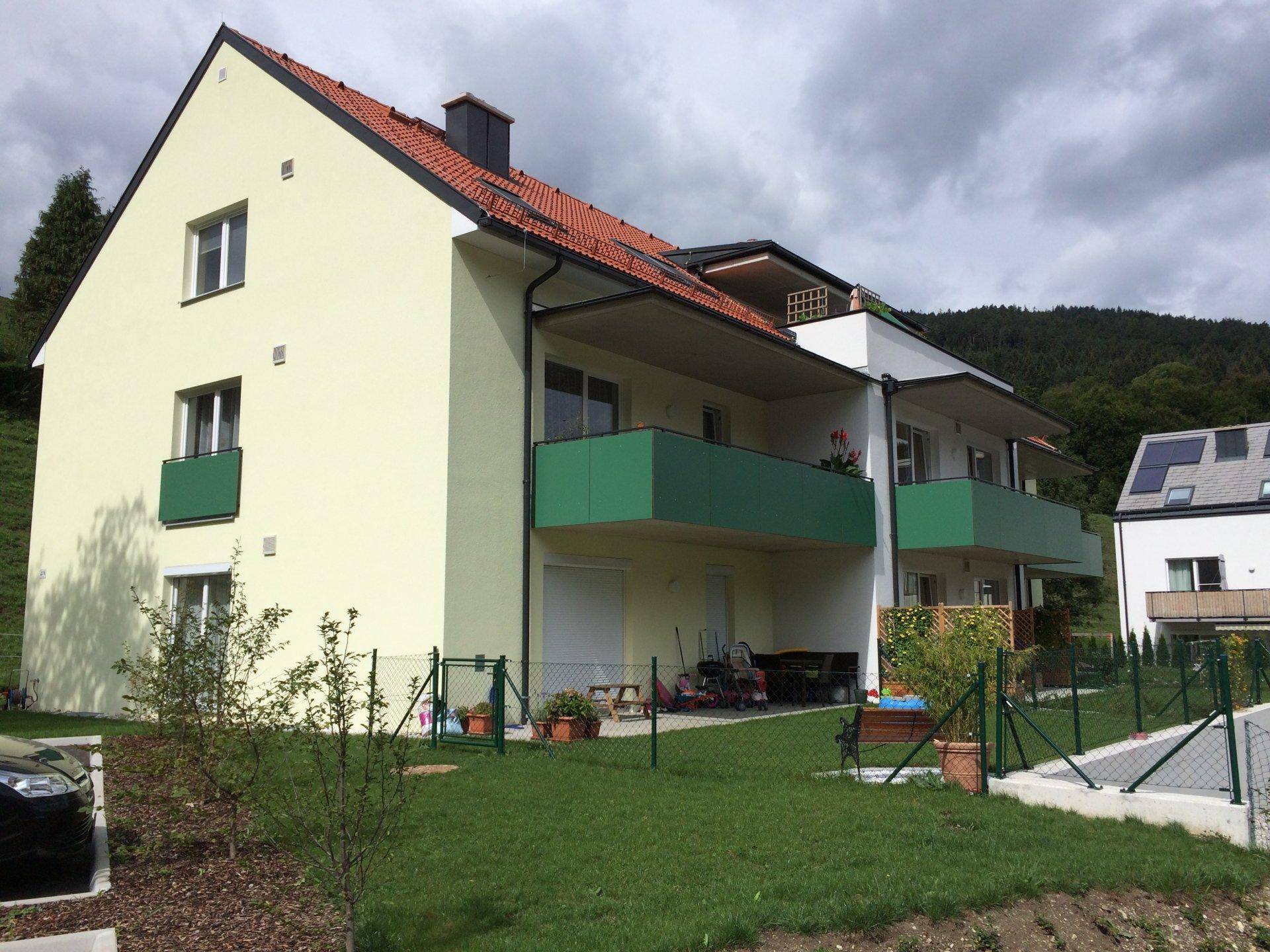 Vorderansicht des Mehrfamilienhauses mit roten Ziegeldächern, grünen Balkonen und eingezäuntem Gartenbereich.