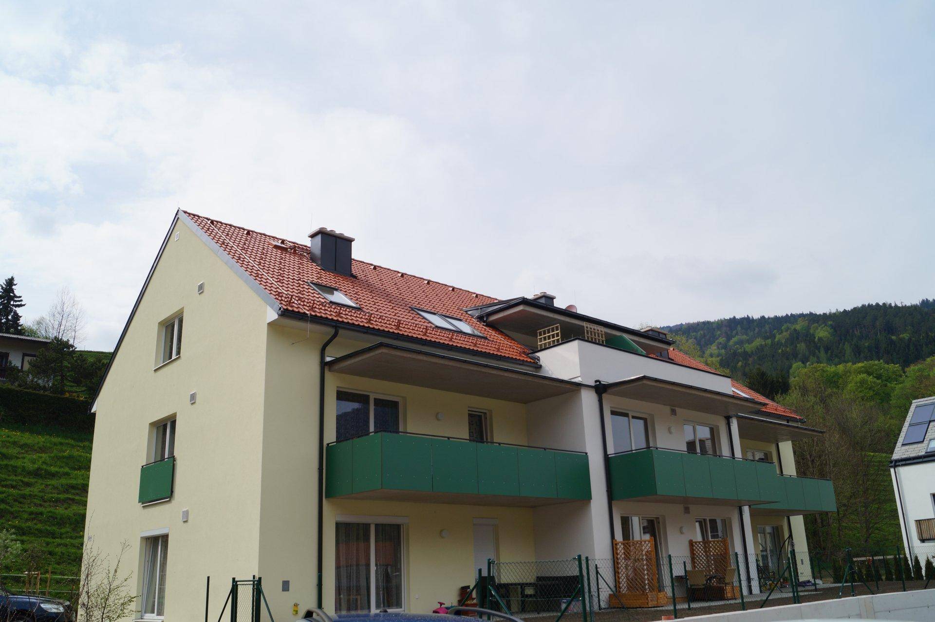 Vorderansicht des Mehrfamilienhauses mit roten Ziegeldächern und grünen Balkonen unter bewölktem Himmel.