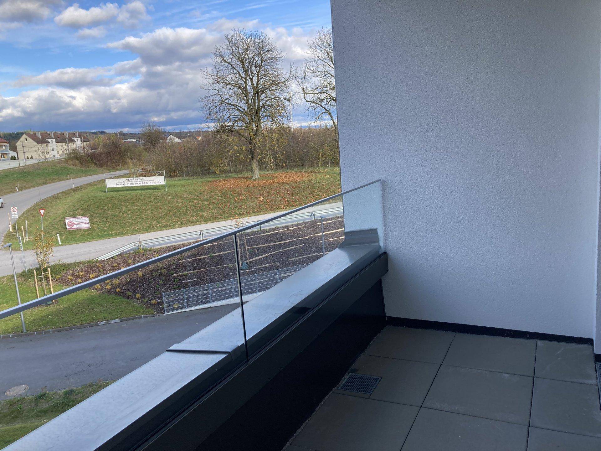 Moderner Balkon mit Glasbrüstung und Blick auf die umliegende Landschaft und Straße unter blauem Himmel.