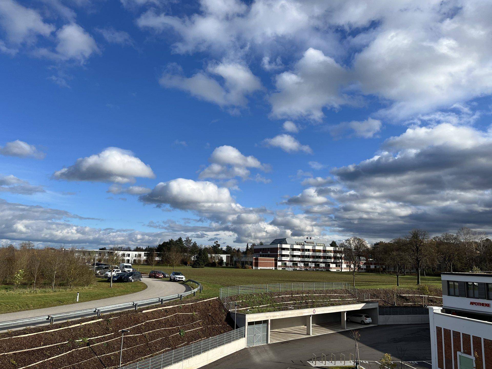 Weitläufiger Blick auf die Umgebung mit Grünflächen, Parkplätzen und weiteren Gebäuden unter blauem Himmel.