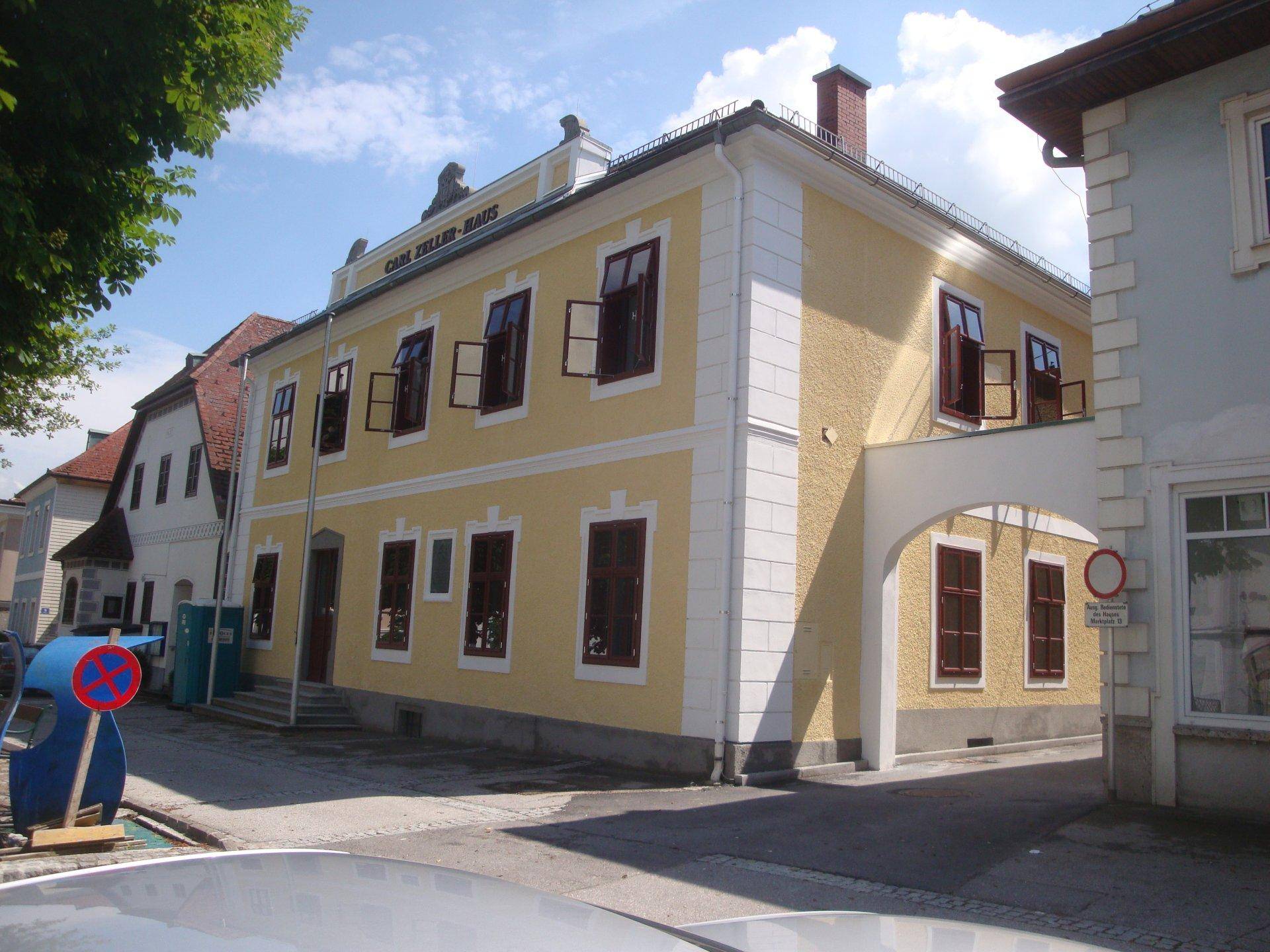 Historisches gelbes Gebäude mit roten Fensterläden und einem Durchgang, umgeben von Bäumen.