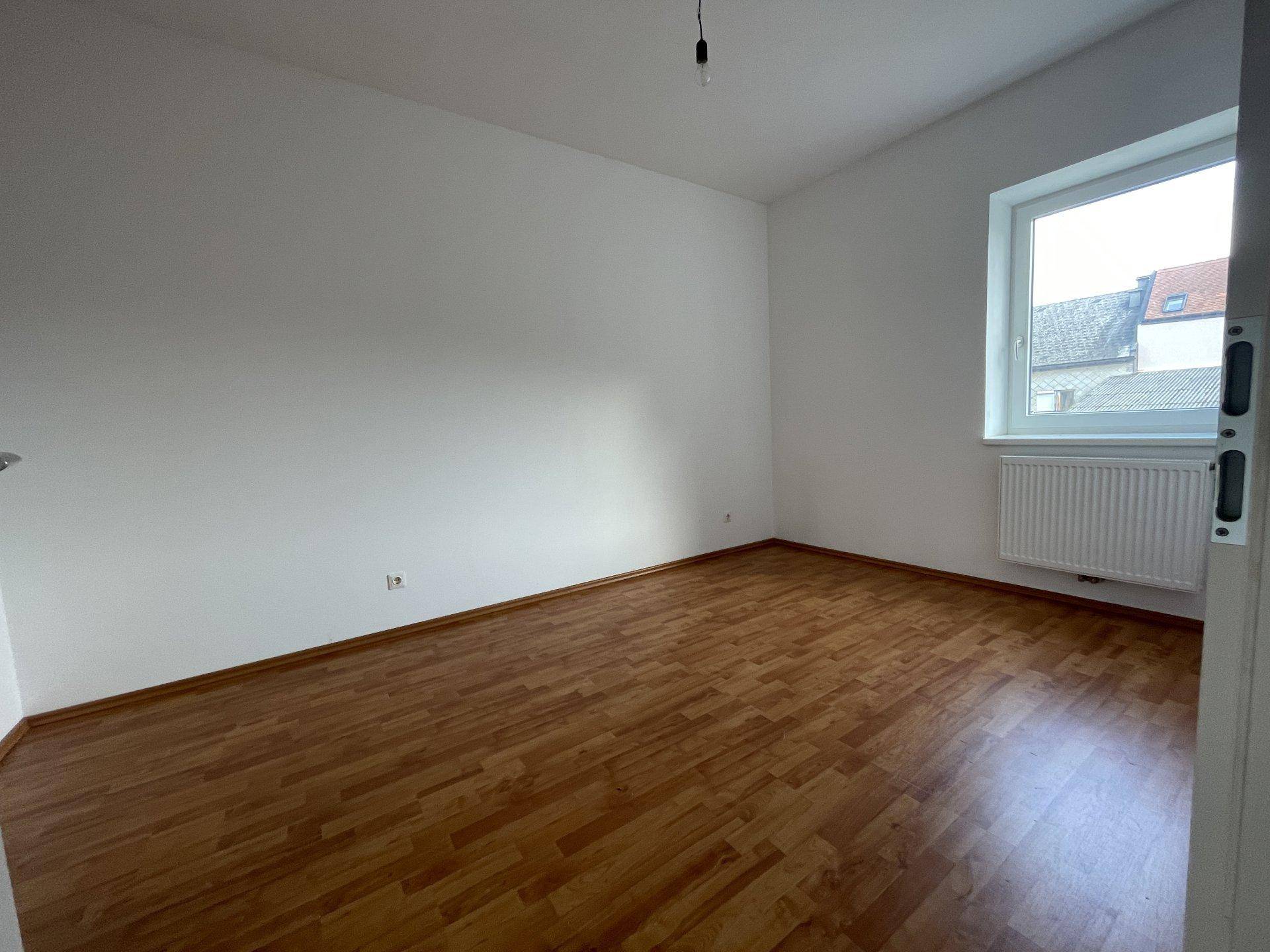 Leeres Schlafzimmer mit Laminatboden, weißen Wänden und einem Fenster mit Blick nach draußen.