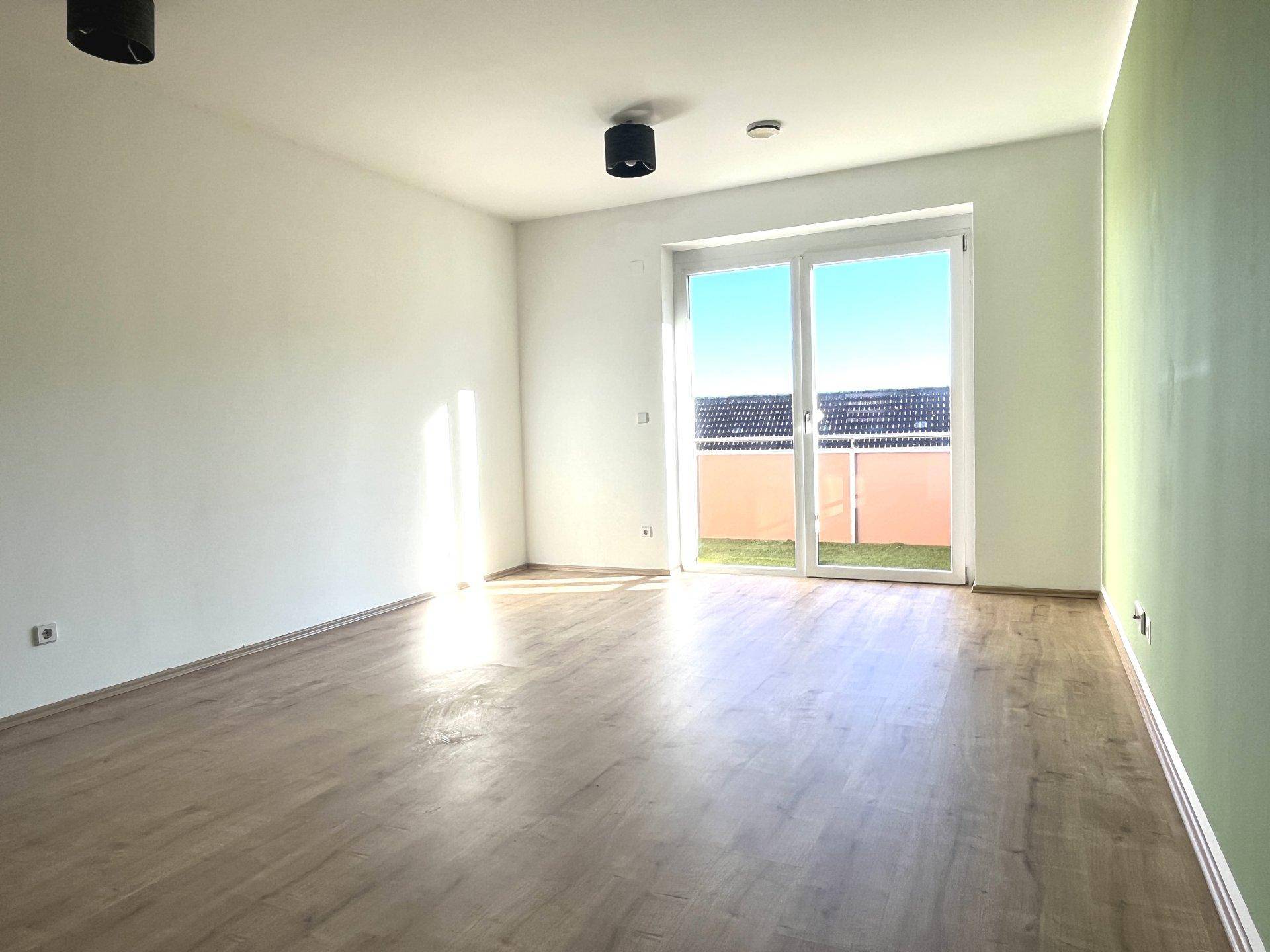 Geräumiges Wohnzimmer mit Laminatboden und großer Terrassentür, die viel Tageslicht hereinlässt.