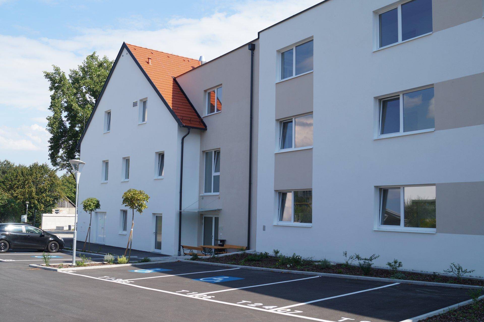 Ansicht der Gebäudefassade mit Parkplätzen und einem traditionellen Dach auf dem angrenzenden Gebäudeteil.