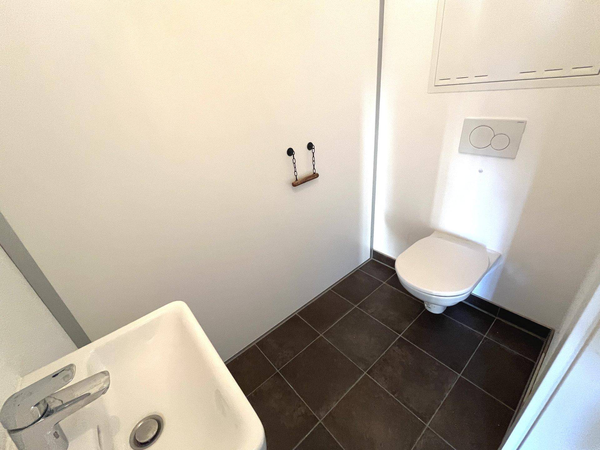 Separates WC mit Wand-WC und kleinem Waschbecken, ausgestattet mit dunklen Bodenfliesen.