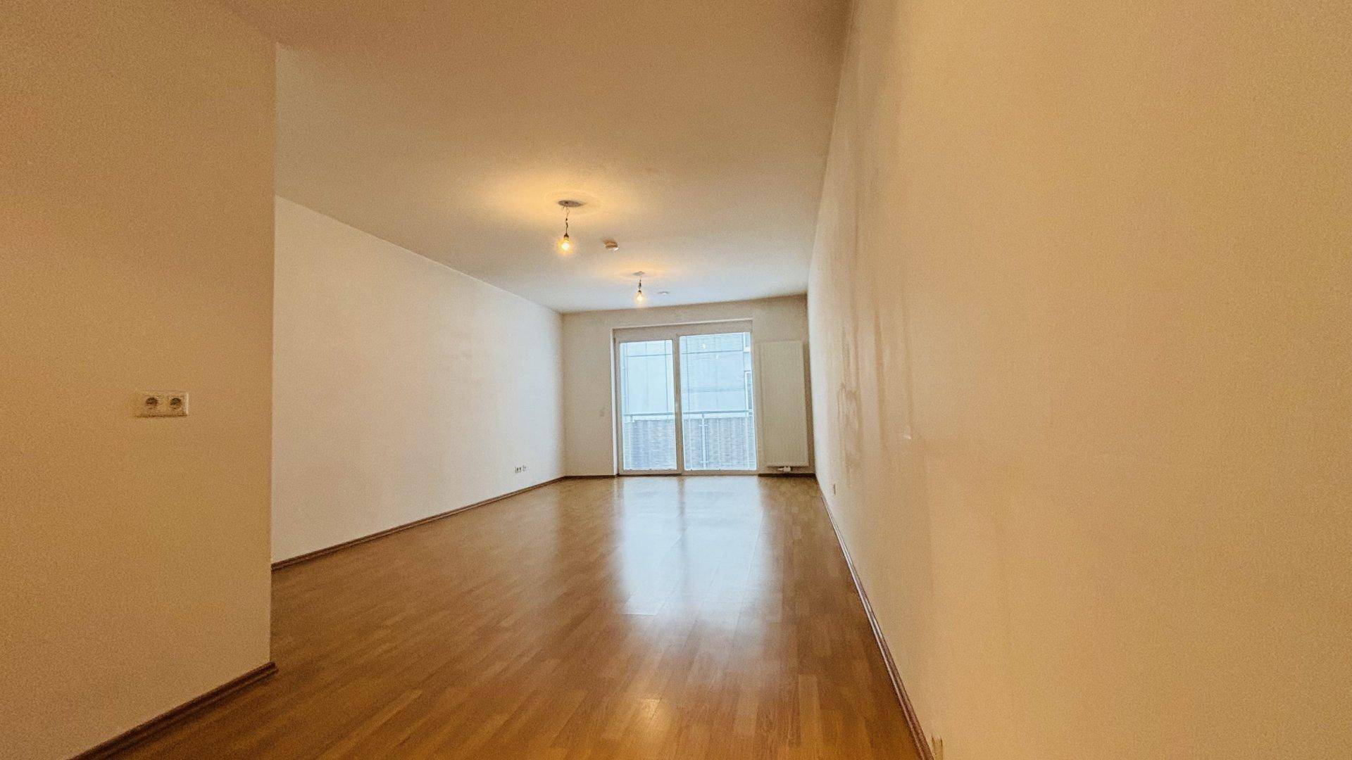 Geräumiger Wohnbereich mit Parkettboden und großem Fenster mit Zugang zu einem Balkon.