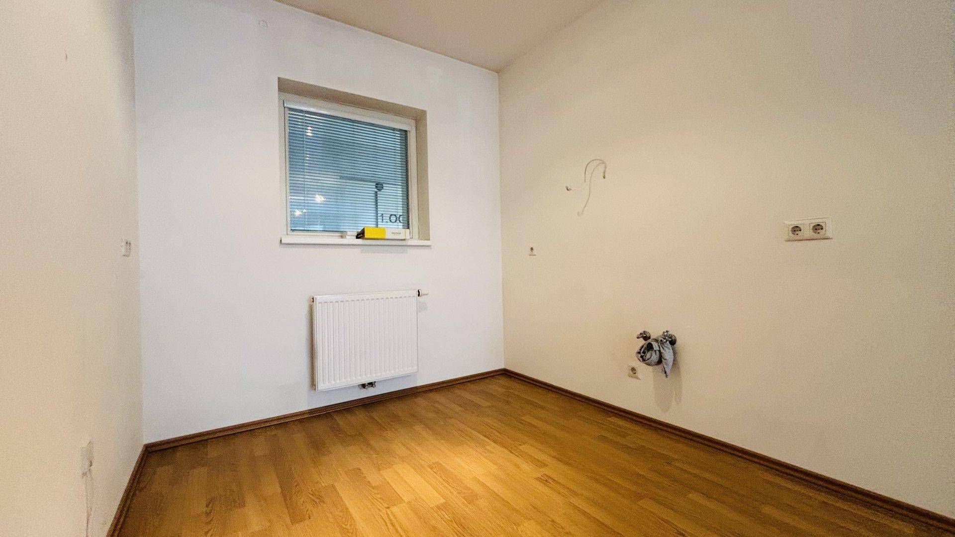 Kleiner Raum mit Parkettboden, einem Fenster und weißen Wänden, vielseitig nutzbar.
