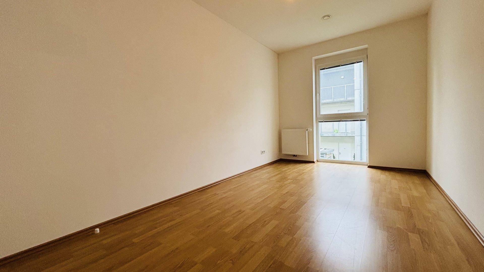 Helles Schlafzimmer mit Parkettboden und großem Fenster, das viel Tageslicht hereinlässt.