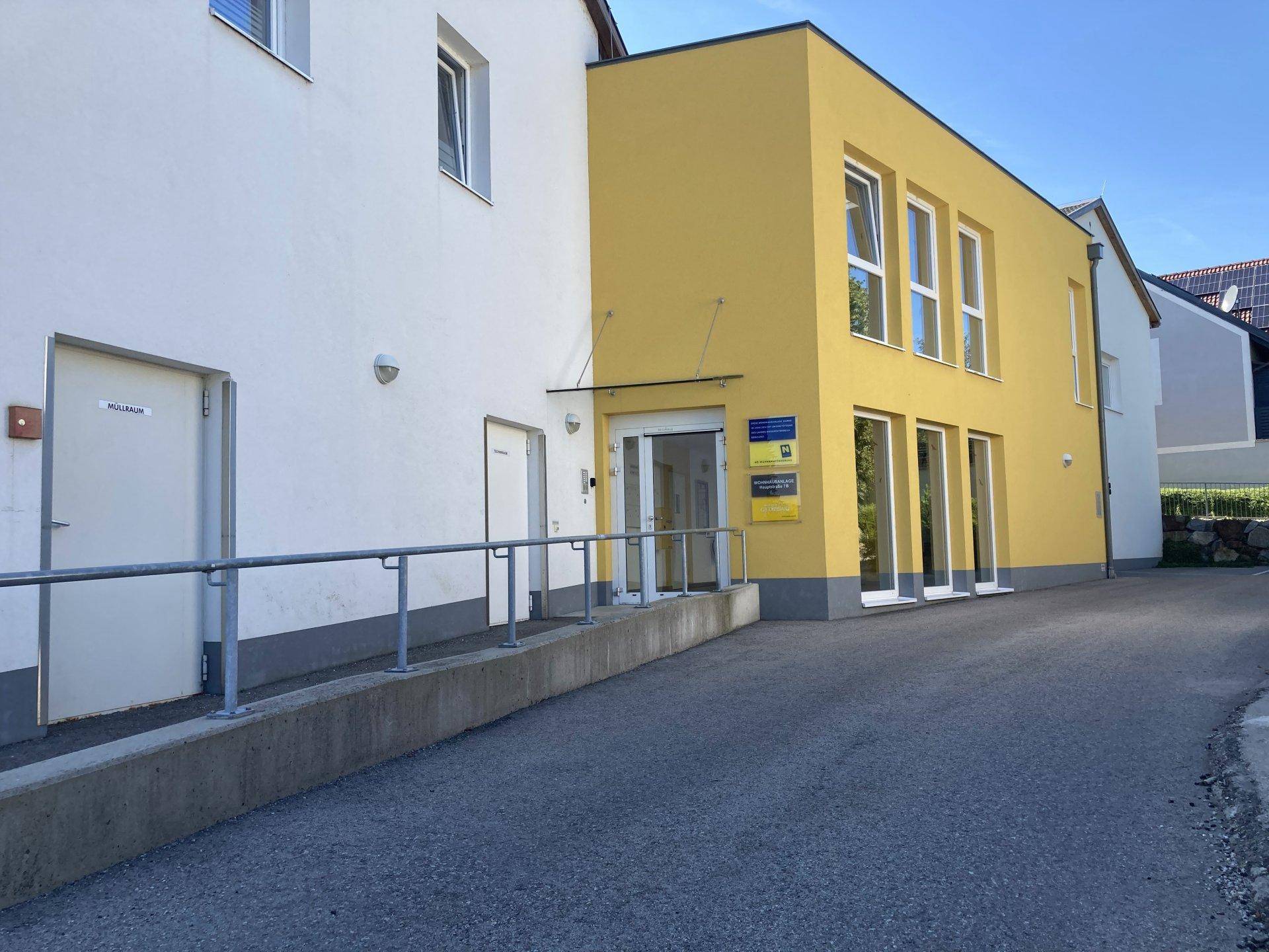 Eingangsbereich der Wohnanlage mit Rampe und großen Fenstern im gelben Gebäudeteil, umgeben von Grün.