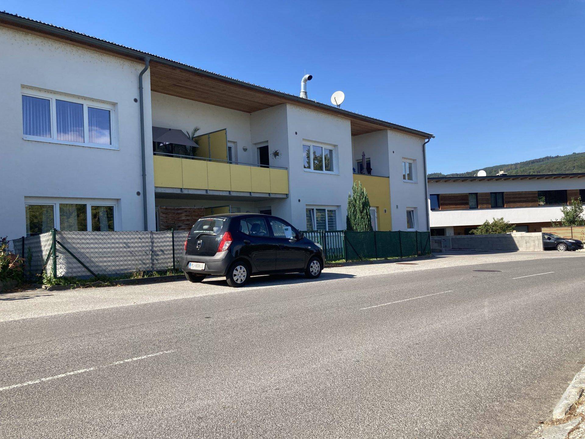 Ansicht der Wohnanlage von der Straße mit Balkonen und geparkten Autos unter strahlendem Himmel.