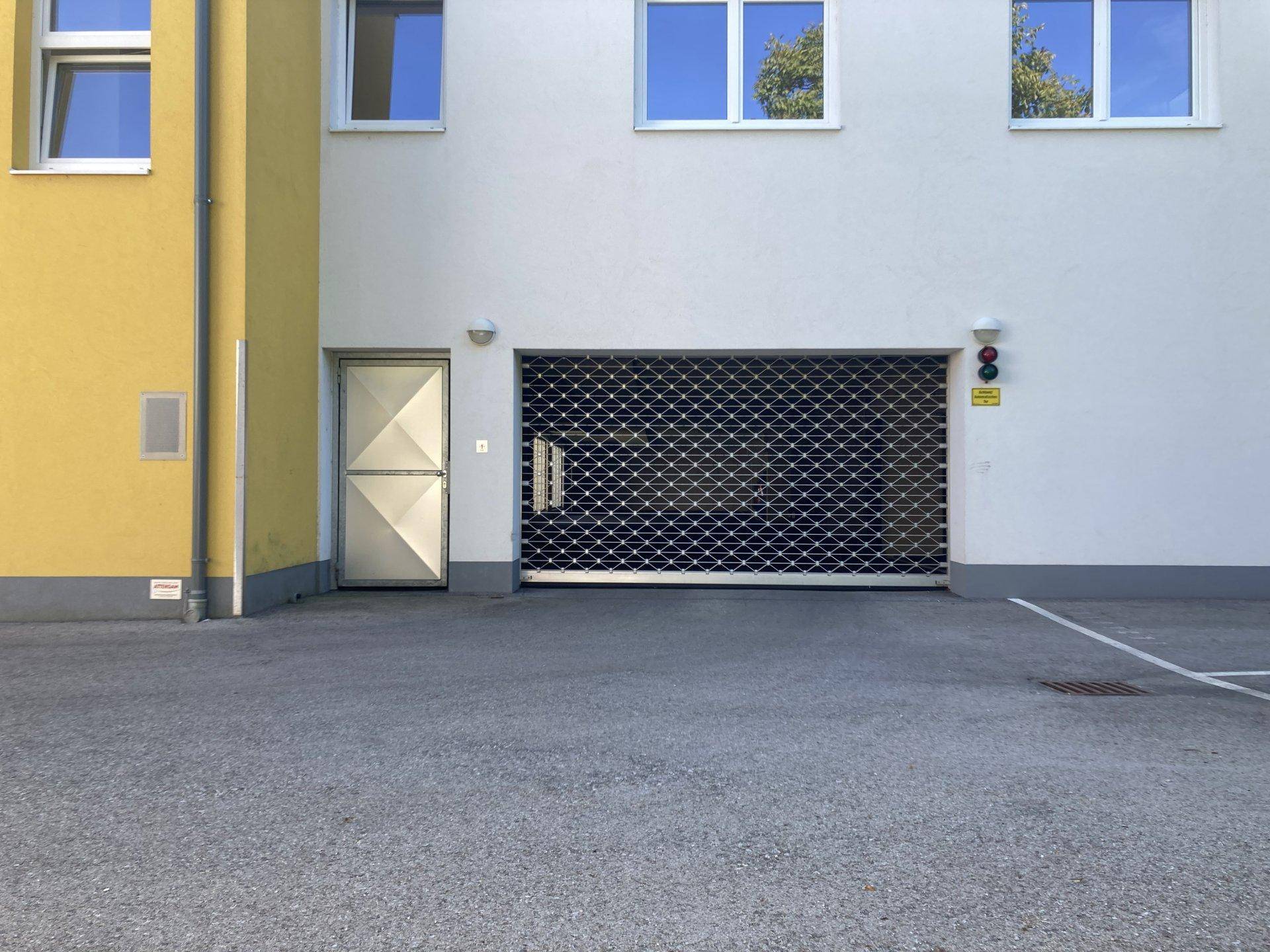 Tiefgarageneinfahrt mit Rolltor und separatem Zugang zur Wohnanlage, umgeben von Asphalt.