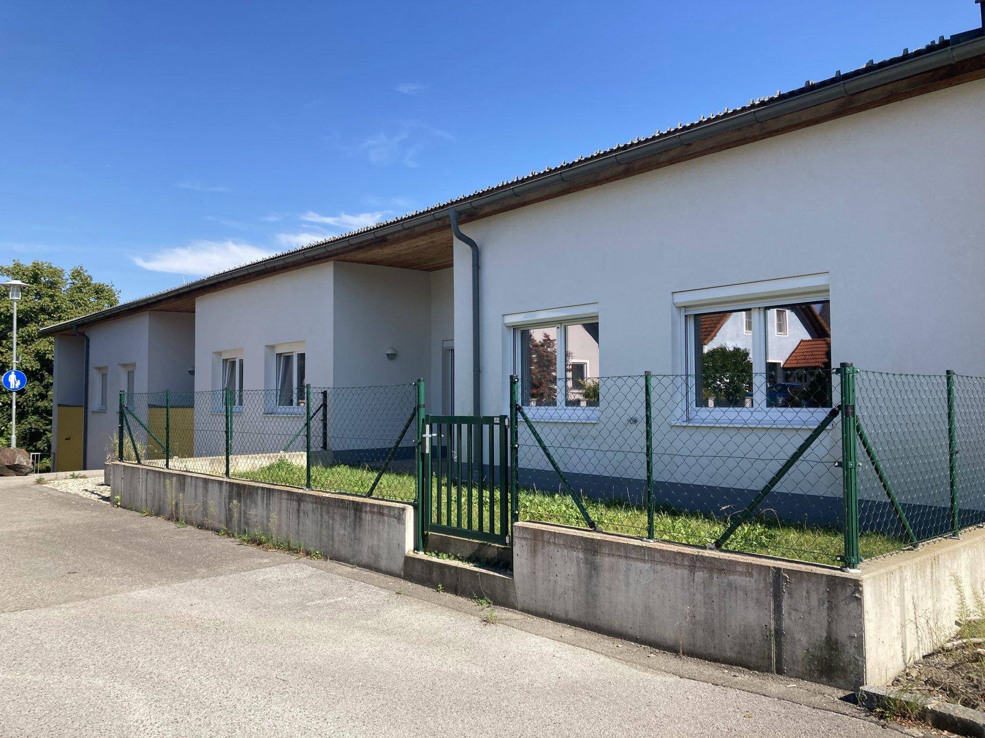 Reihenhausähnliche Wohngebäude mit kleinen Vorgärten und Rollläden an den Fenstern unter blauem Himmel.