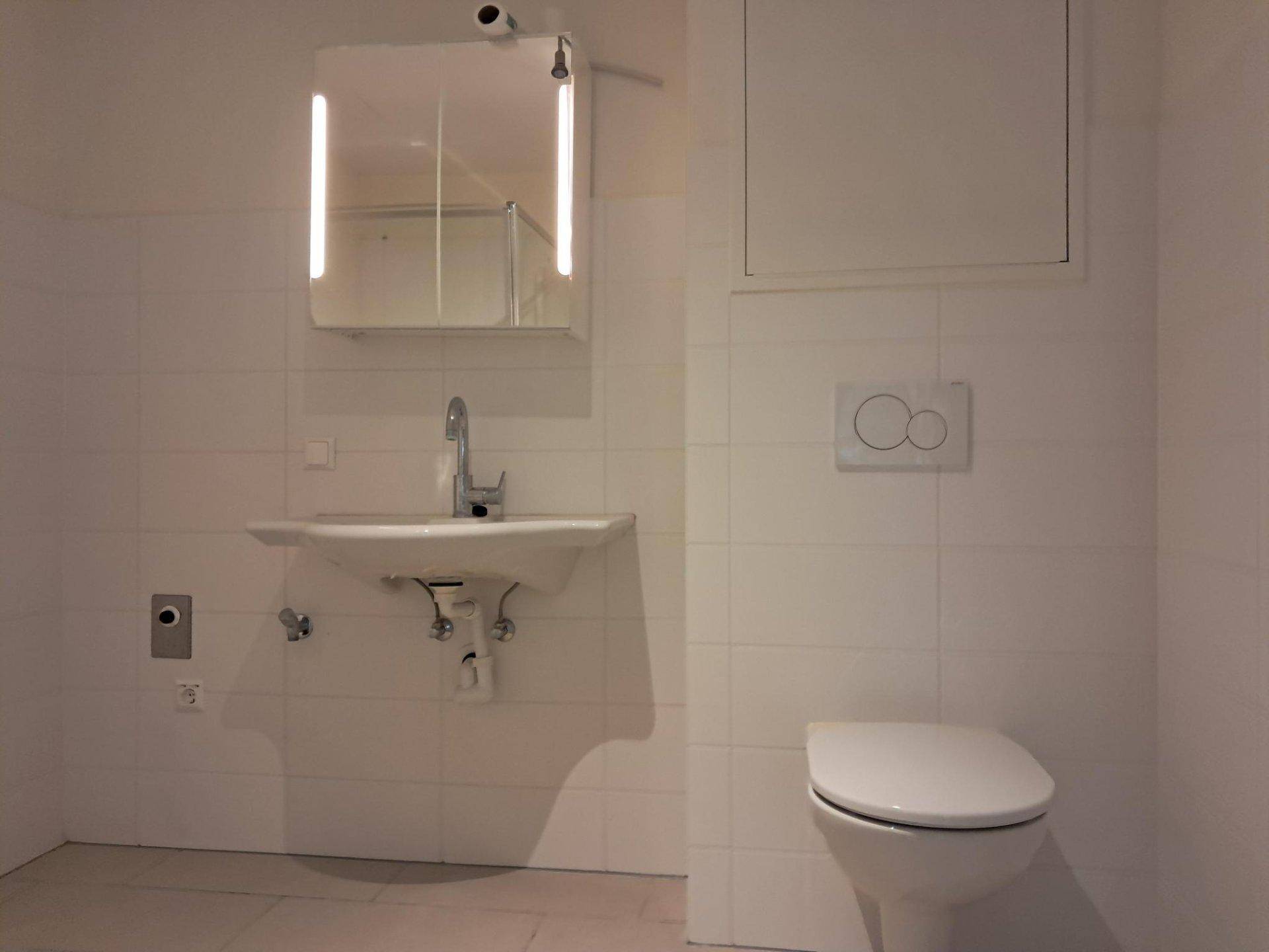 Modernes Badezimmer mit Wand-WC, Waschbecken und beleuchtetem Spiegelschrank in hellen Fliesen.