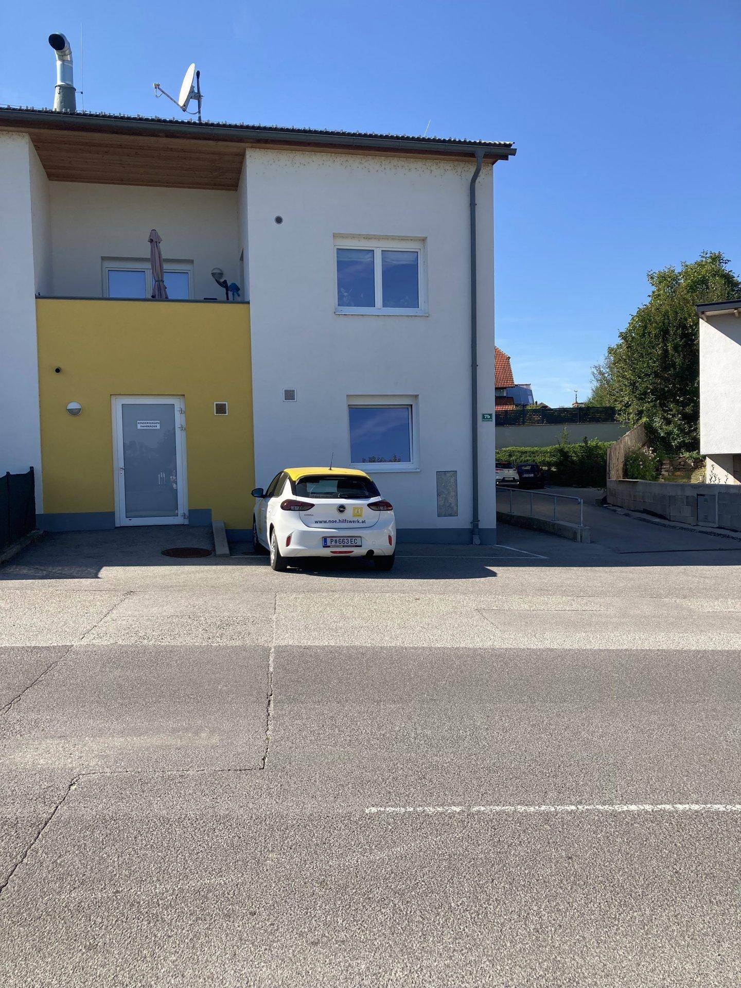 Vorderansicht der Wohnanlage mit Parkplatz und einem geparkten Fahrzeug unter blauem Himmel.