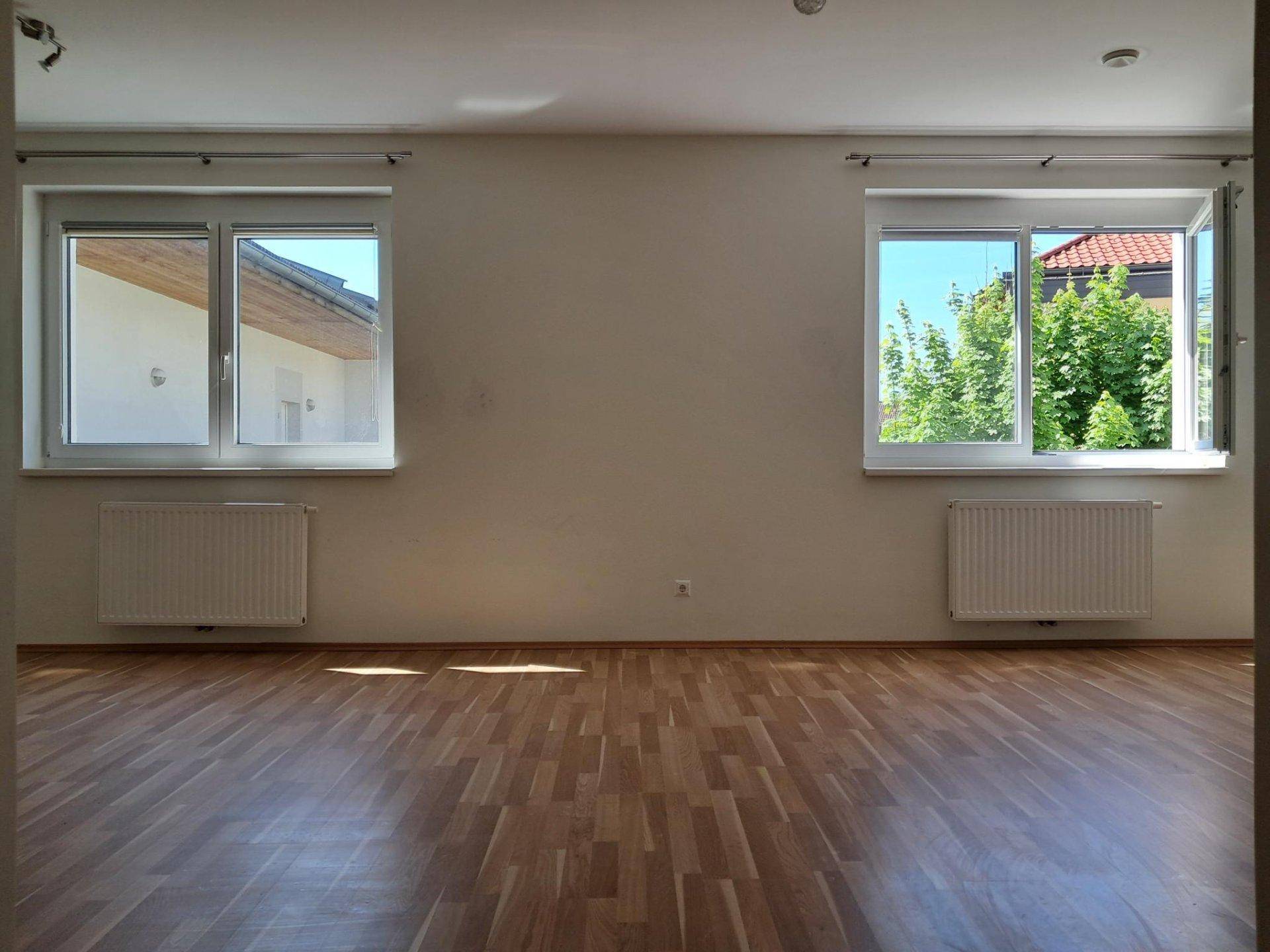Heller Wohnraum mit Laminatboden und zwei Fenstern, die viel Tageslicht hereinlassen.
