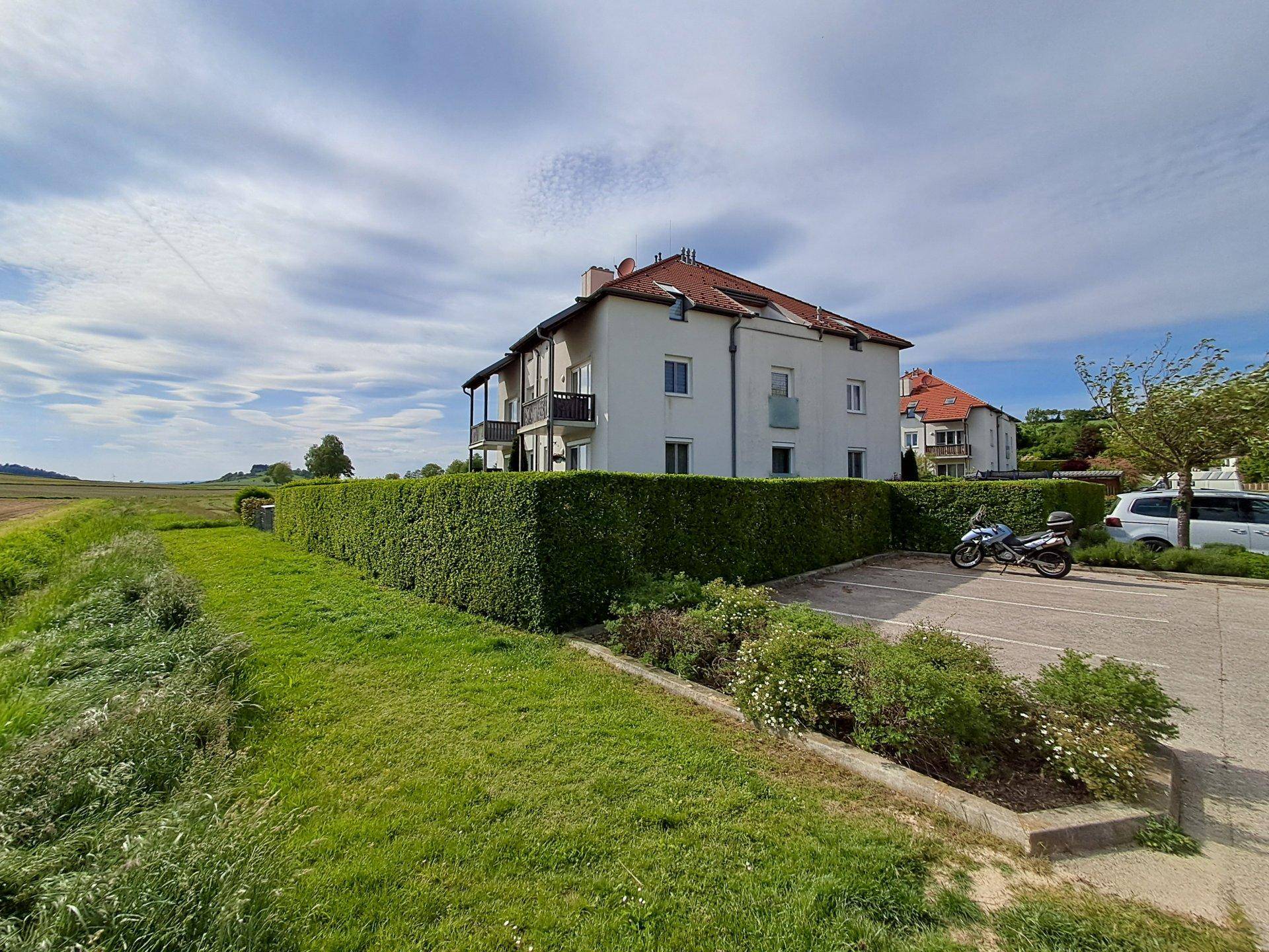 Rückansicht des Gebäudes mit Parkplätzen, hoher Hecke und weitem Blick auf die Landschaft.
