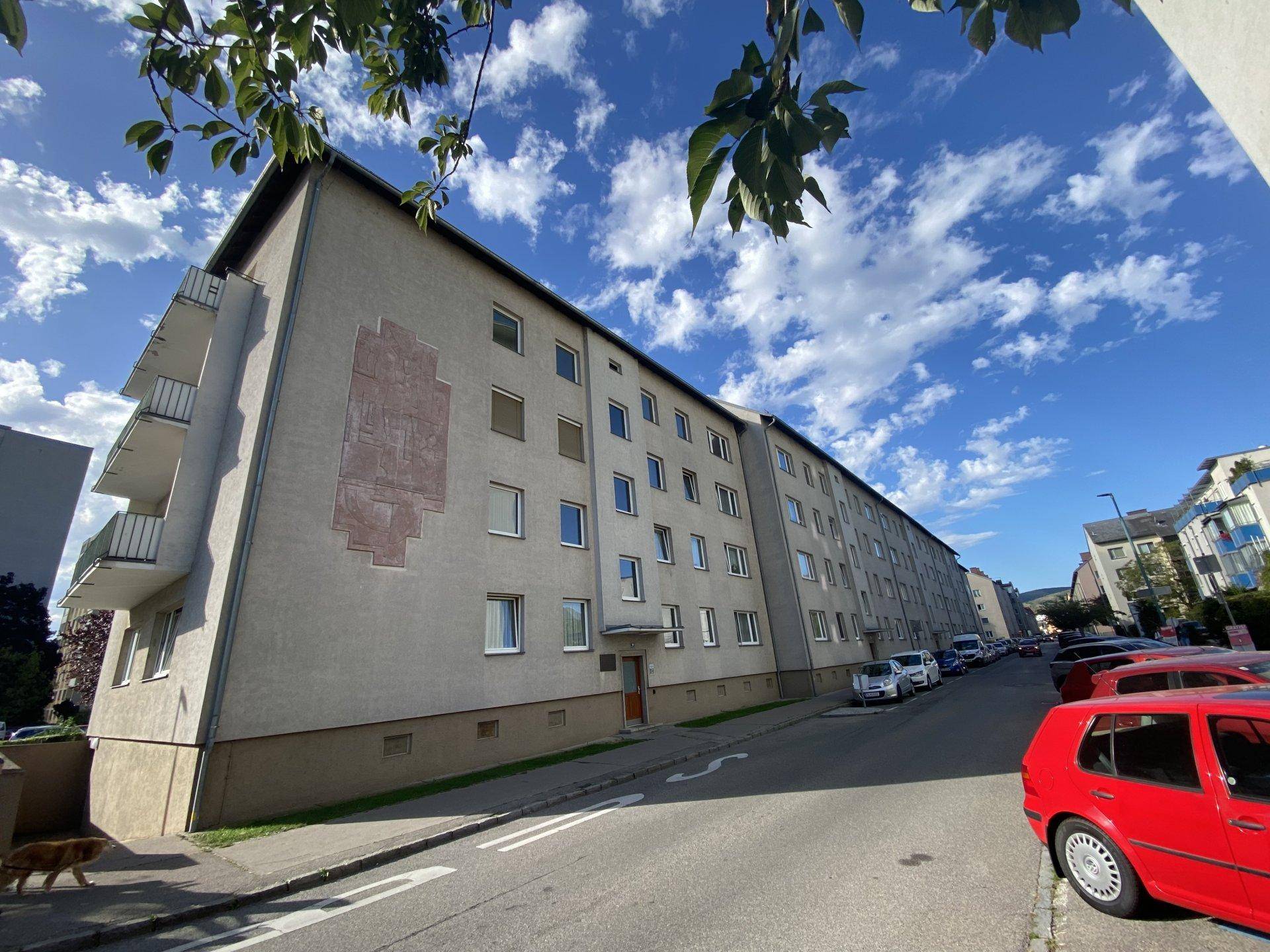 Lange Fassade eines Wohngebäudes entlang einer Straße mit geparkten Autos an einem sonnigen Tag.