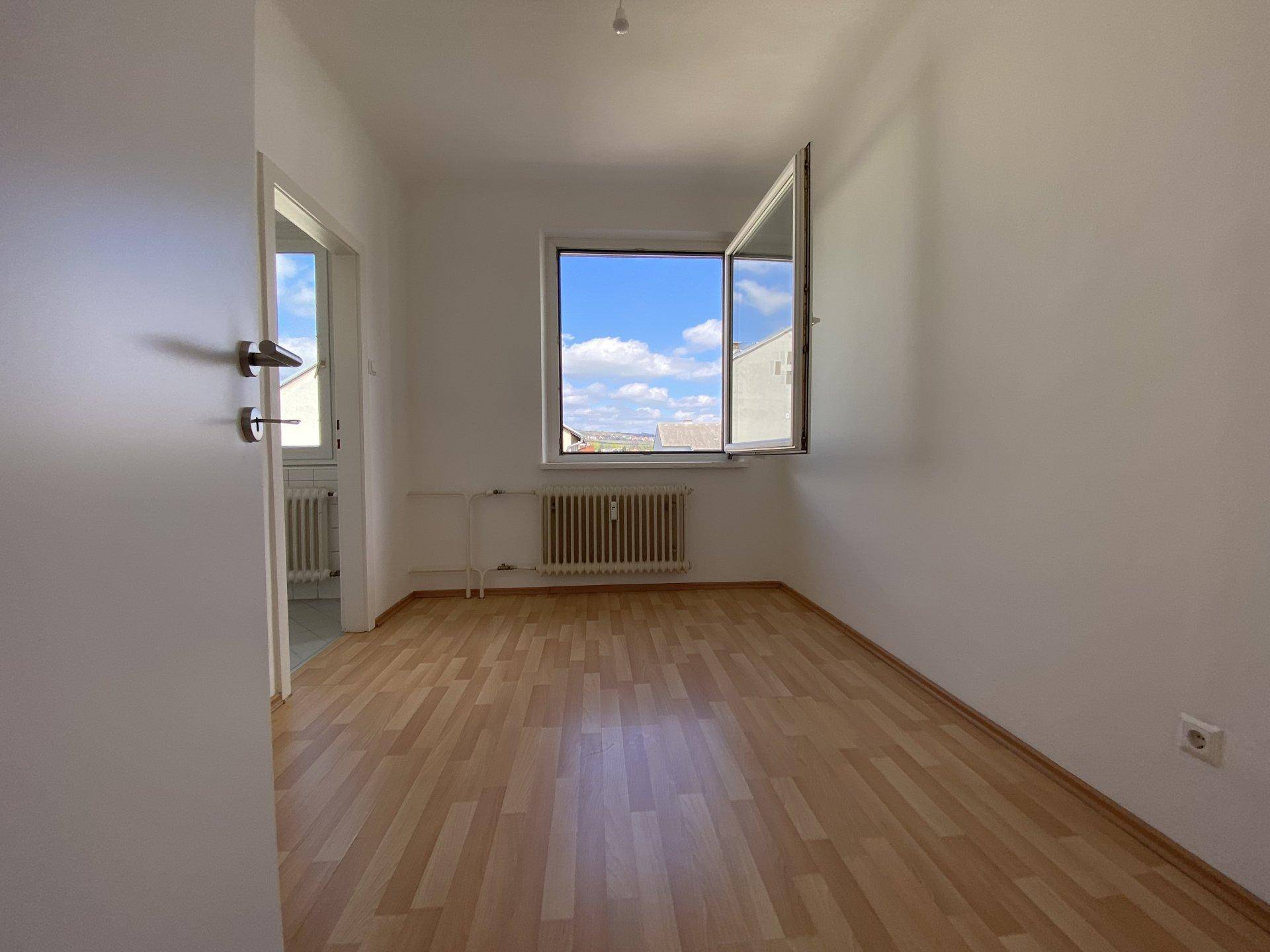 Leeres Zimmer mit hellem Laminatboden, weißen Wänden und einem geöffneten Fenster mit Blick nach draußen.