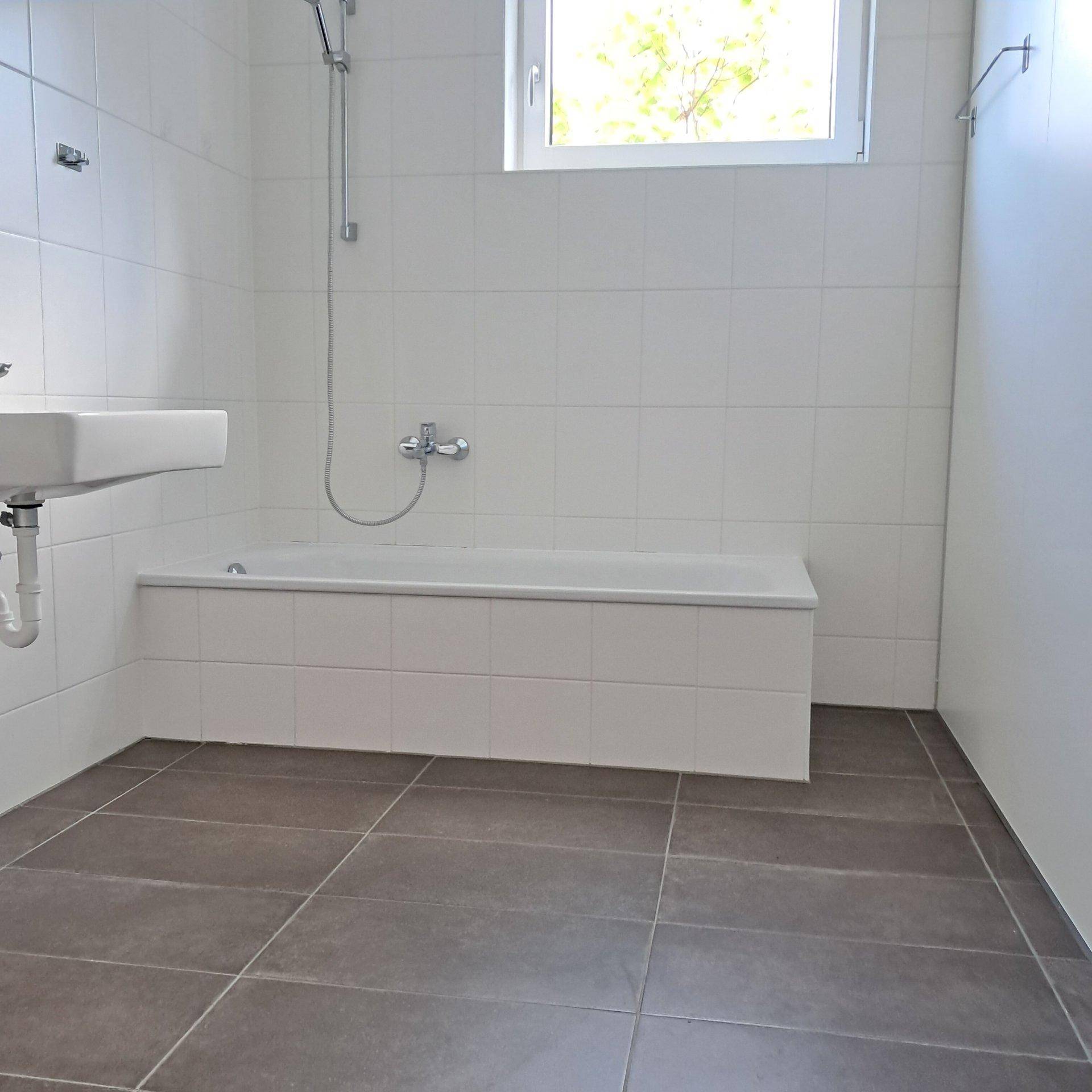 Helles Badezimmer mit Badewanne, Waschbecken und Fenster, komplett gefliest.