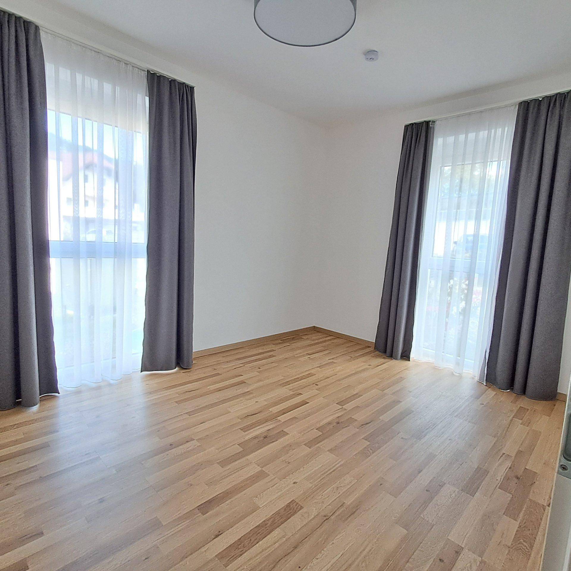Helles Schlafzimmer mit Holzboden und zwei großen Fenstern mit Vorhängen.