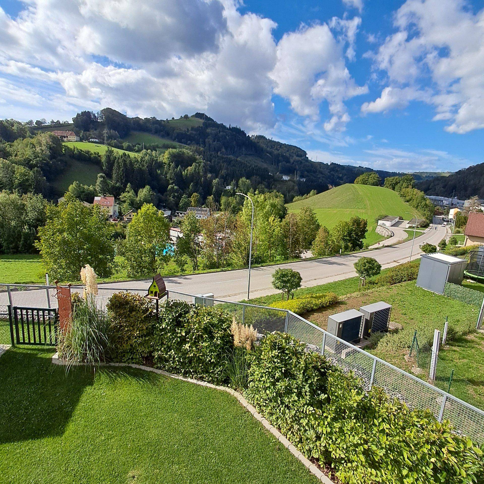 Weitläufiger Blick auf die grüne Landschaft mit Hügeln und einer Straße unter blauem Himmel.