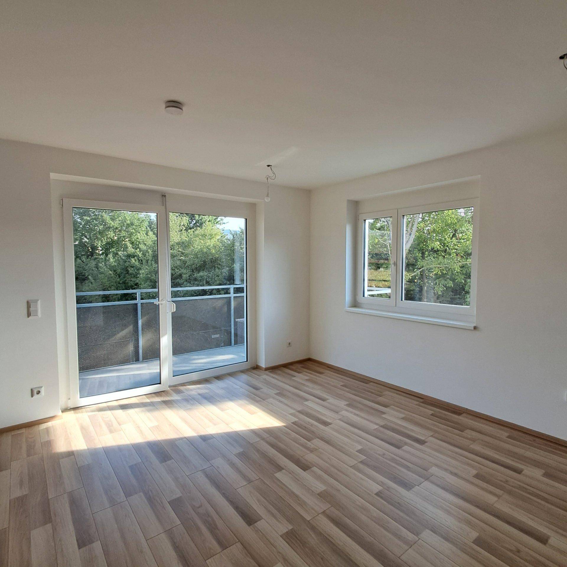 Heller Raum mit Laminatboden, Balkonzugang und großen Fenstern mit Blick ins Grüne.