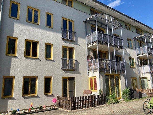 Seitenansicht des Wohngebäudes mit Balkonen, gelben Fensterrahmen und kleiner Zaunbegrenzung.