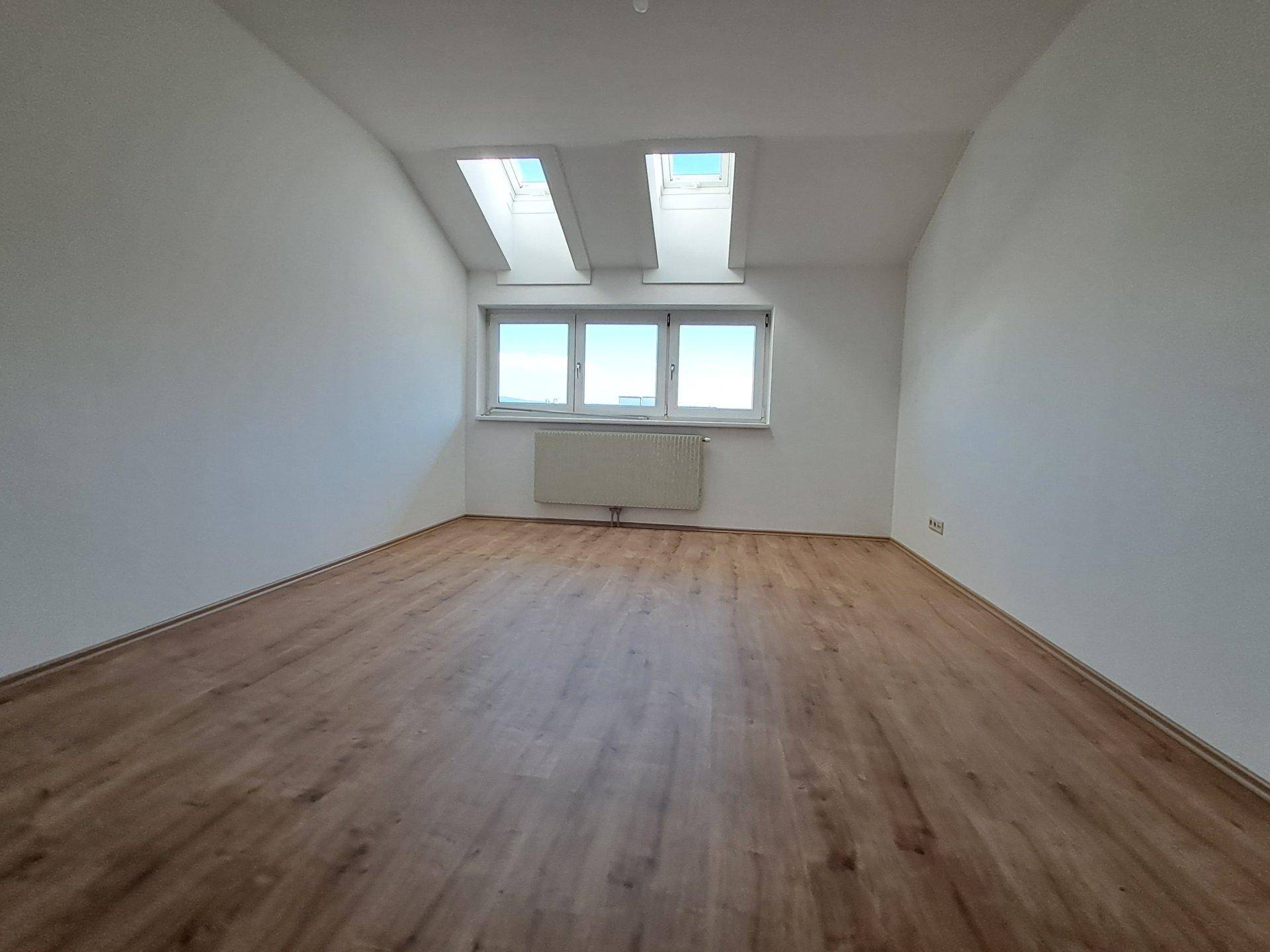 Lichtdurchflutetes Zimmer mit Laminatboden, großen Fenstern und zwei Dachfenstern.