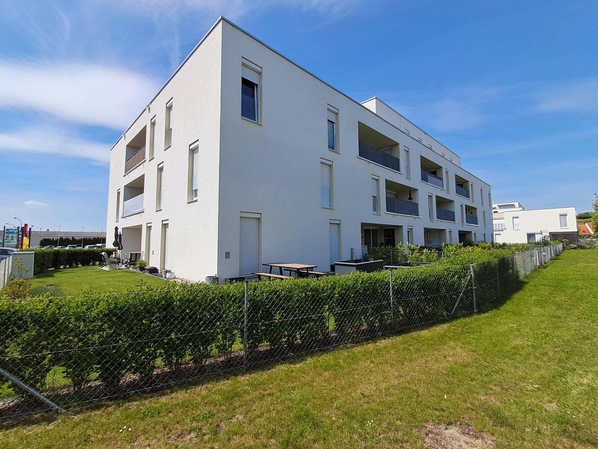 Seitenansicht eines modernen Apartmentgebäudes mit weißer Fassade und umlaufender Hecke.