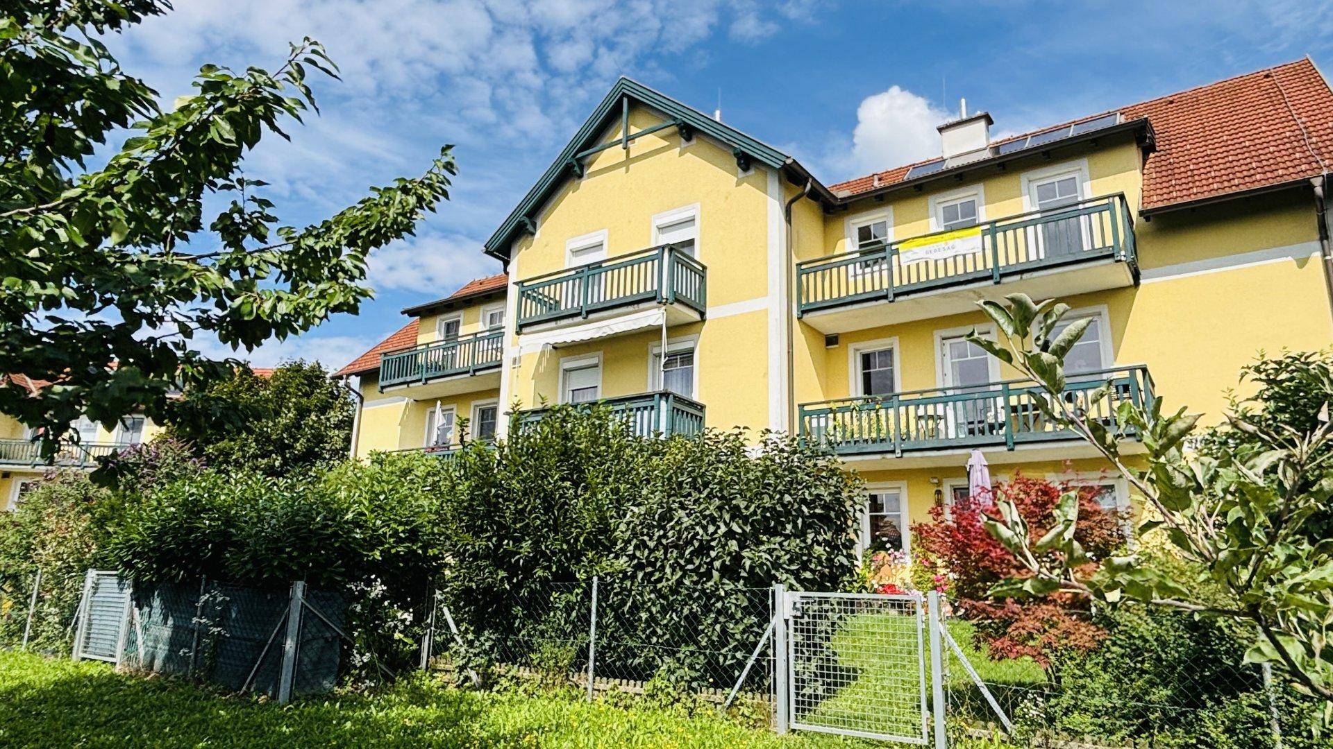 Fassade des gelben Gebäudes mit Balkonen, umgeben von gepflegten Hecken und Bäumen unter blauem Himmel.