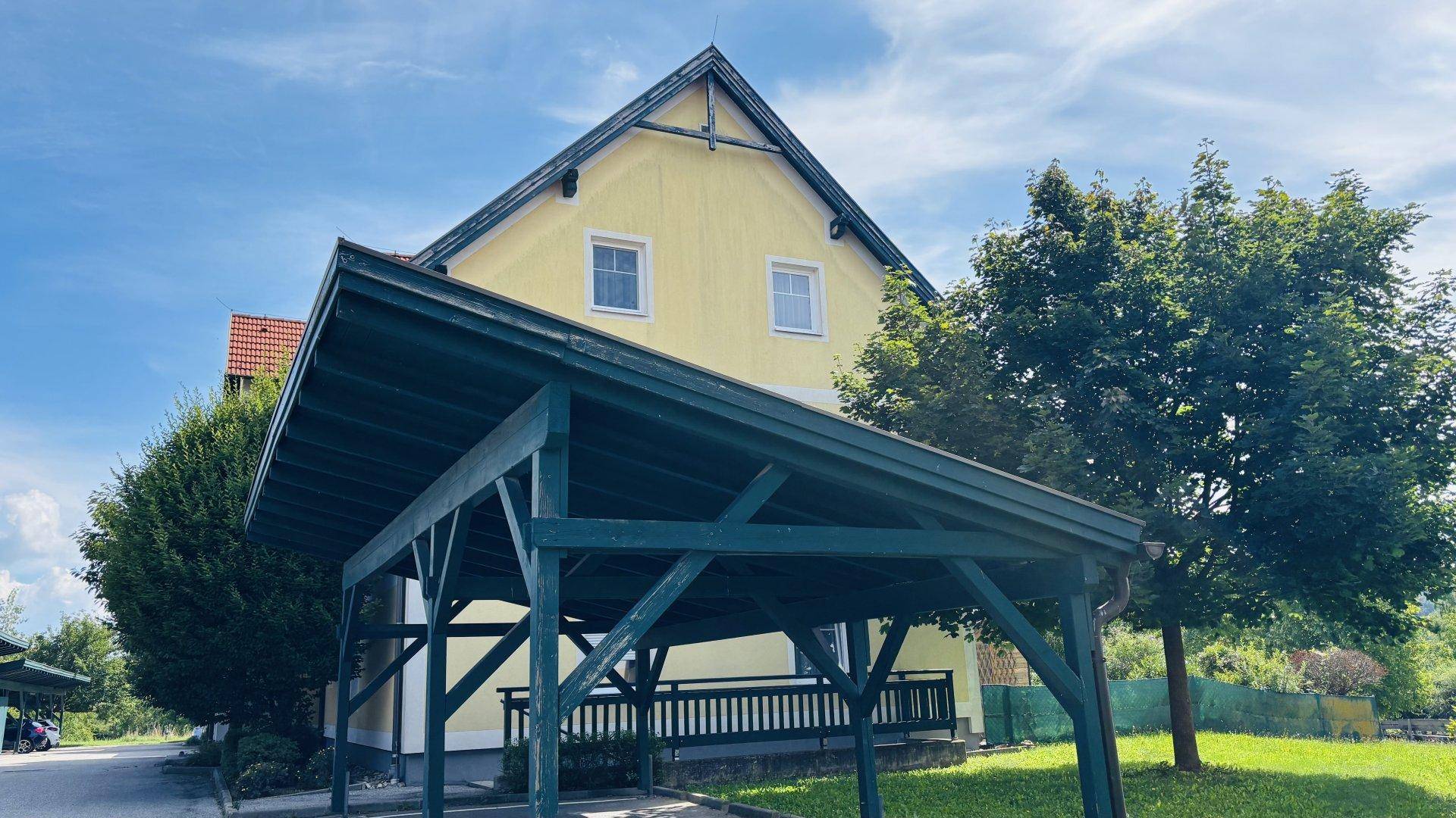 Seitenansicht des Gebäudes mit einem hölzernen Carport und grüner Umgebung unter blauem Himmel.