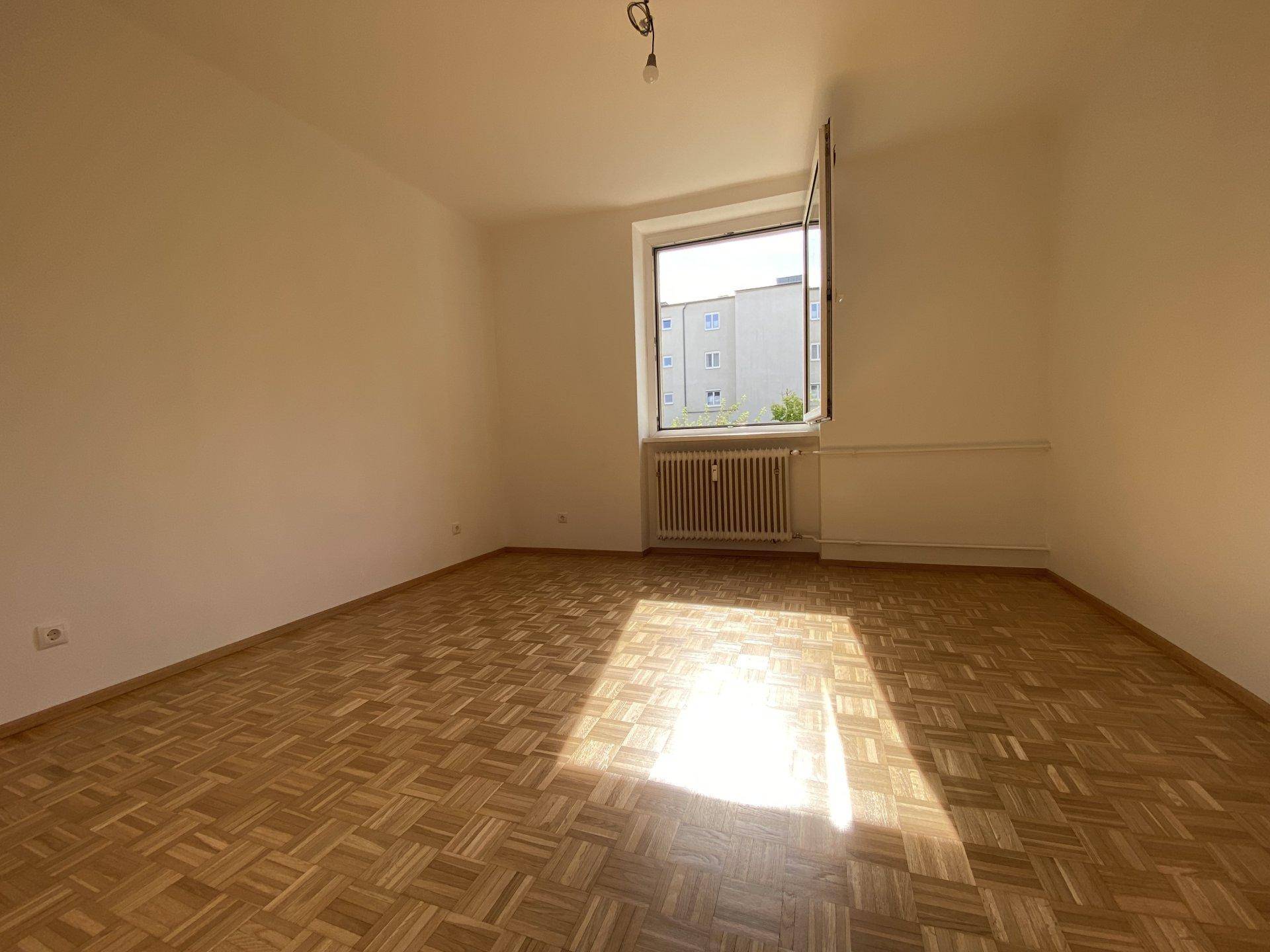 Helles Schlafzimmer mit klassischem Parkettboden und einem Fenster, das viel Sonnenlicht hereinlässt.