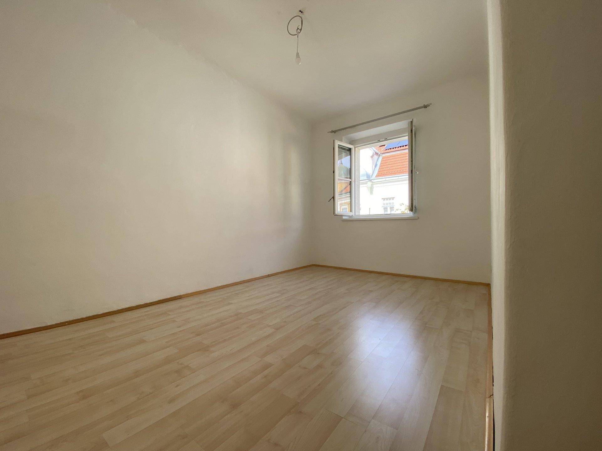 Helles, leeres Zimmer mit Laminatboden und einem geöffneten Fenster, das Tageslicht hereinlässt.