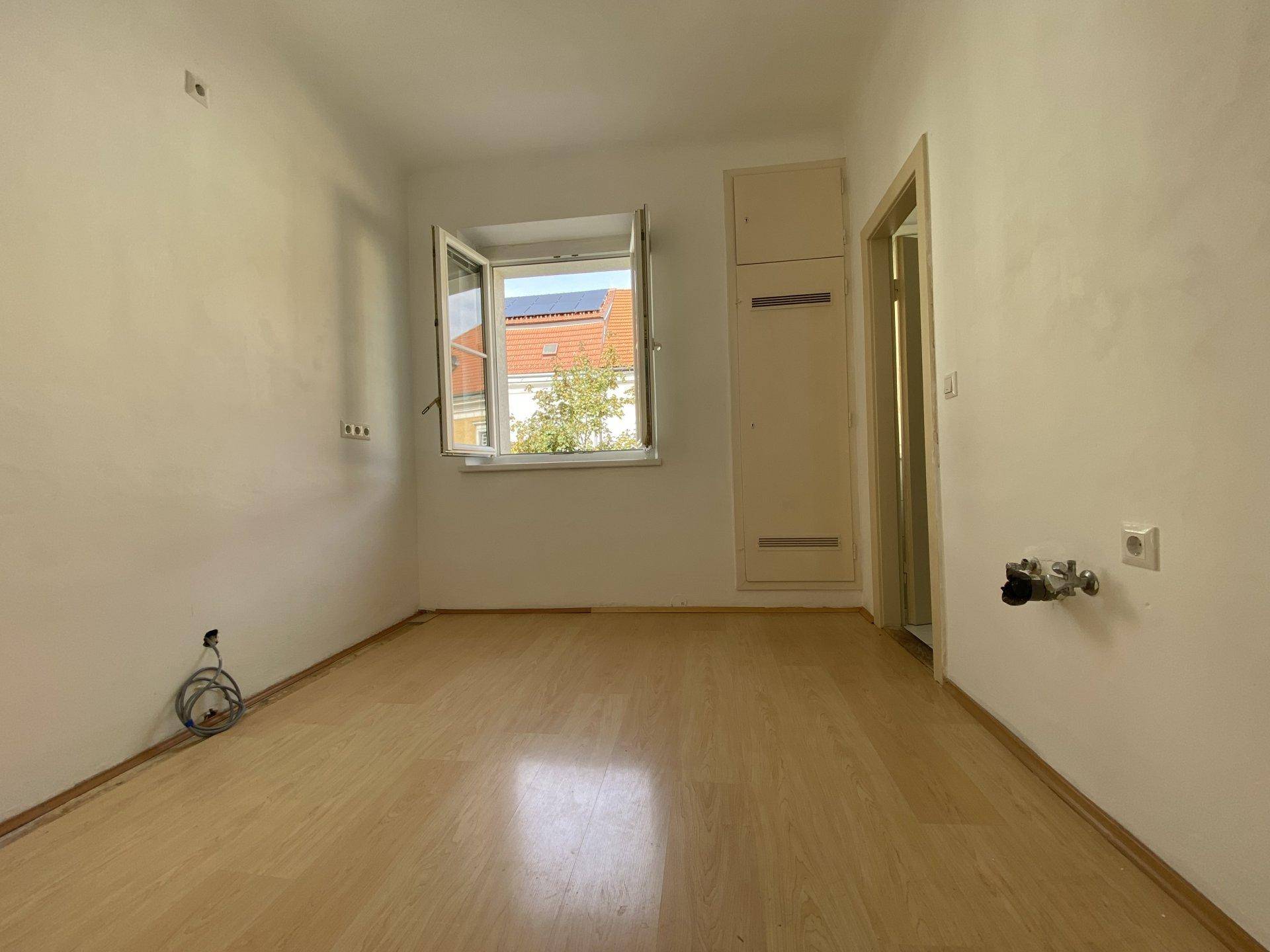 Leerer Raum mit Fenster, hellem Laminatboden und Anschlüssen für eine zukünftige Küche.