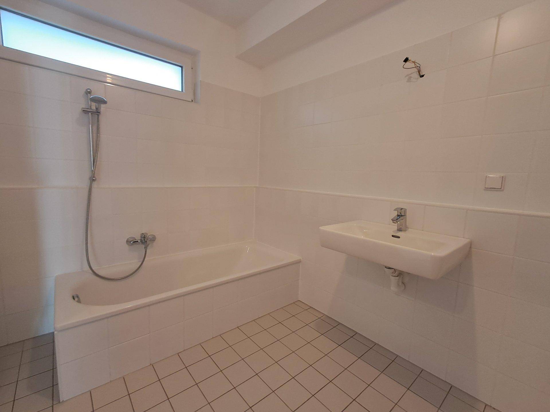 Helles Badezimmer mit Badewanne, Duscharmatur, Waschbecken und Fenster.
