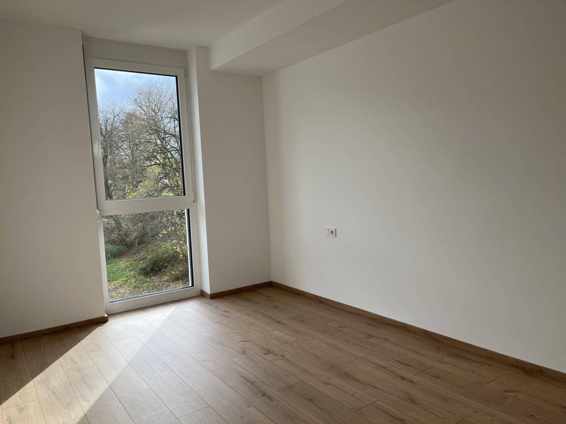 Lichtdurchflutetes Zimmer mit Laminatboden und großem Fenster, das viel Tageslicht hereinlässt.