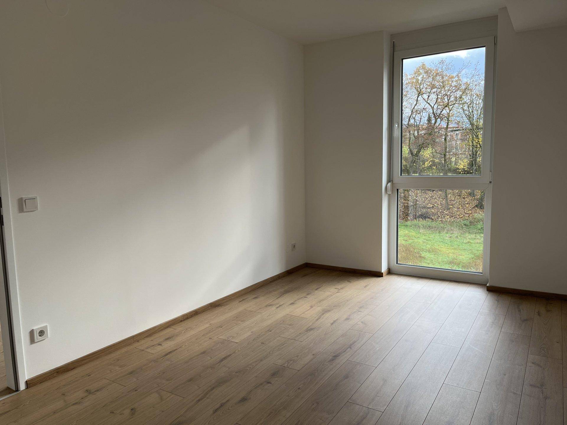 Helles Schlafzimmer mit Laminatboden und großem Fenster mit Blick ins Grüne.
