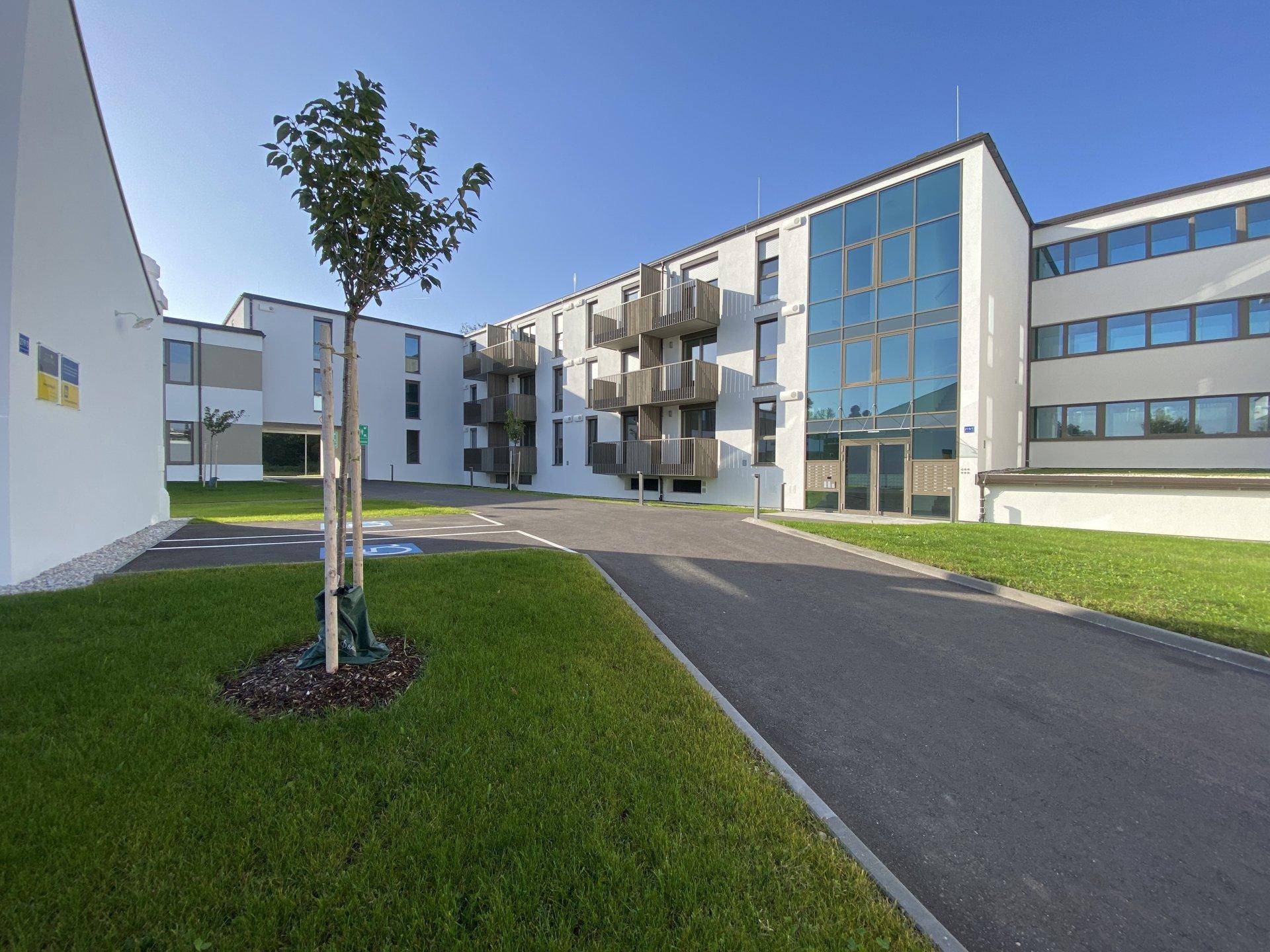 Ansicht eines modernen Wohnkomplexes mit Glasfassade am Eingang und Zufahrtsweg.