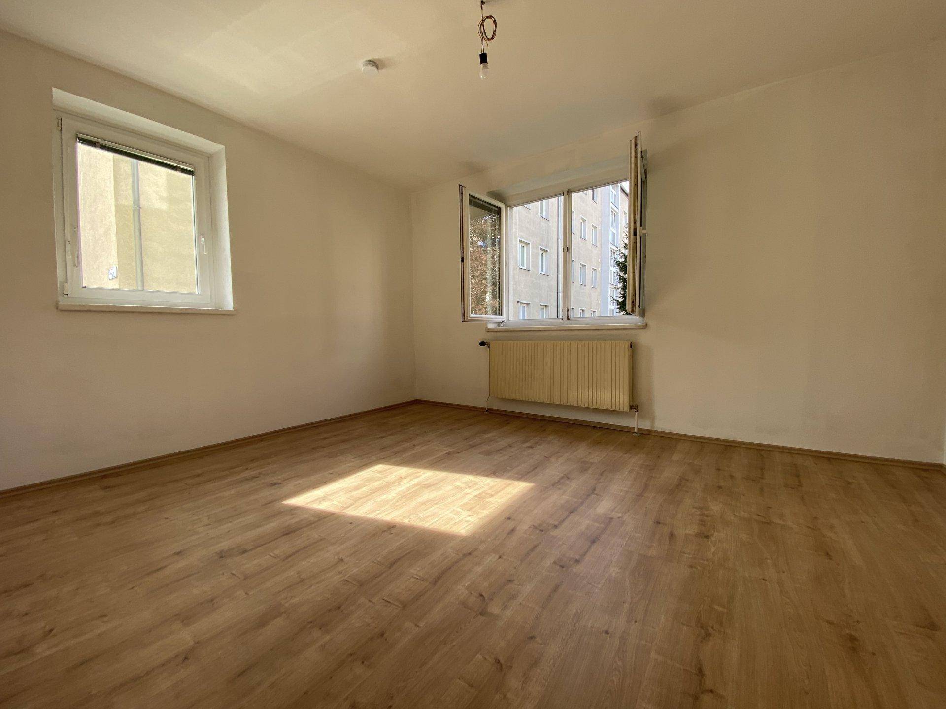 Helles Wohnzimmer mit Laminatboden, zwei Fenstern und Sonneneinfall.